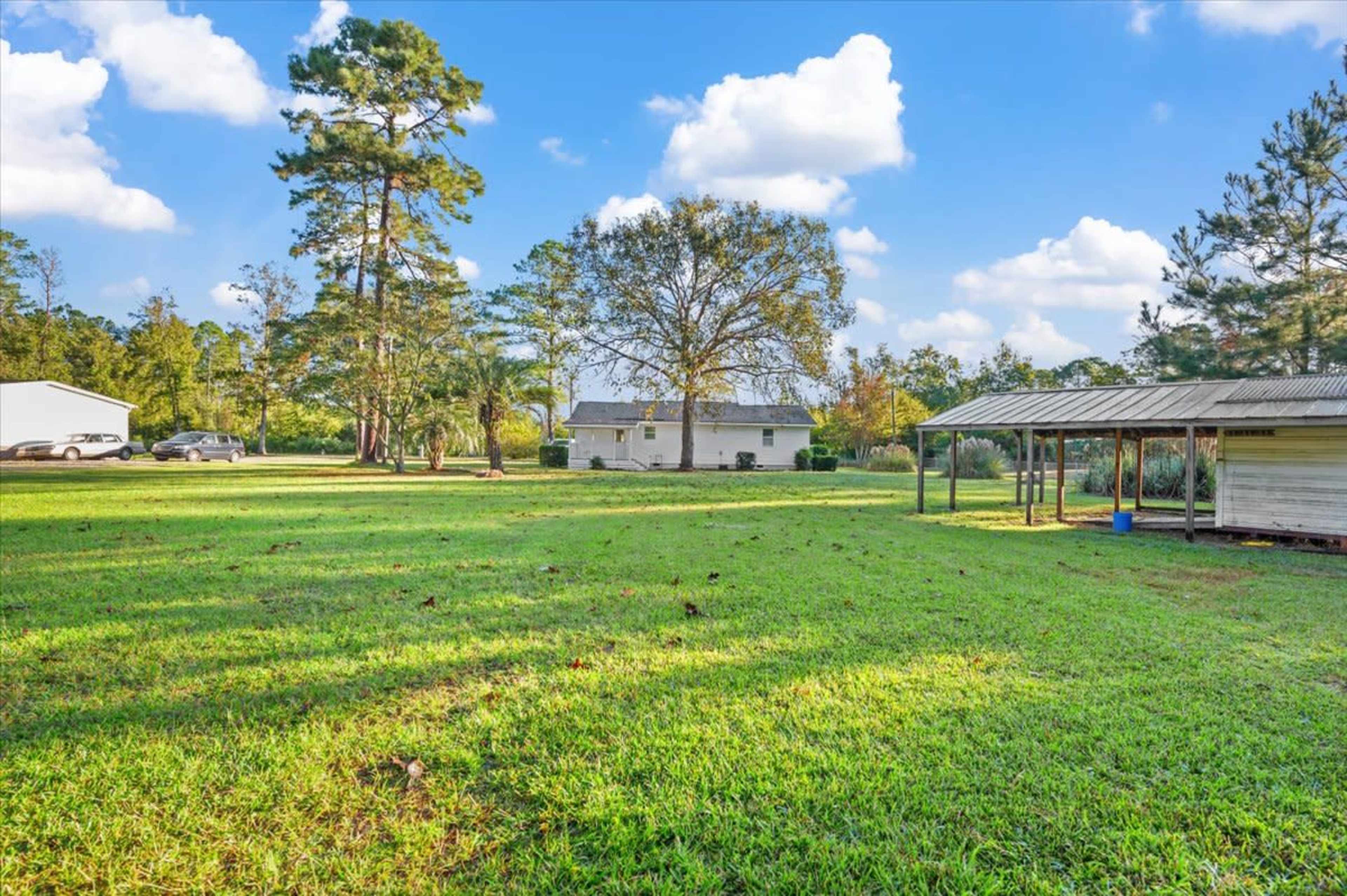 A spacious yard with a house in the background, surrounded by trees and a covered structure on one side.