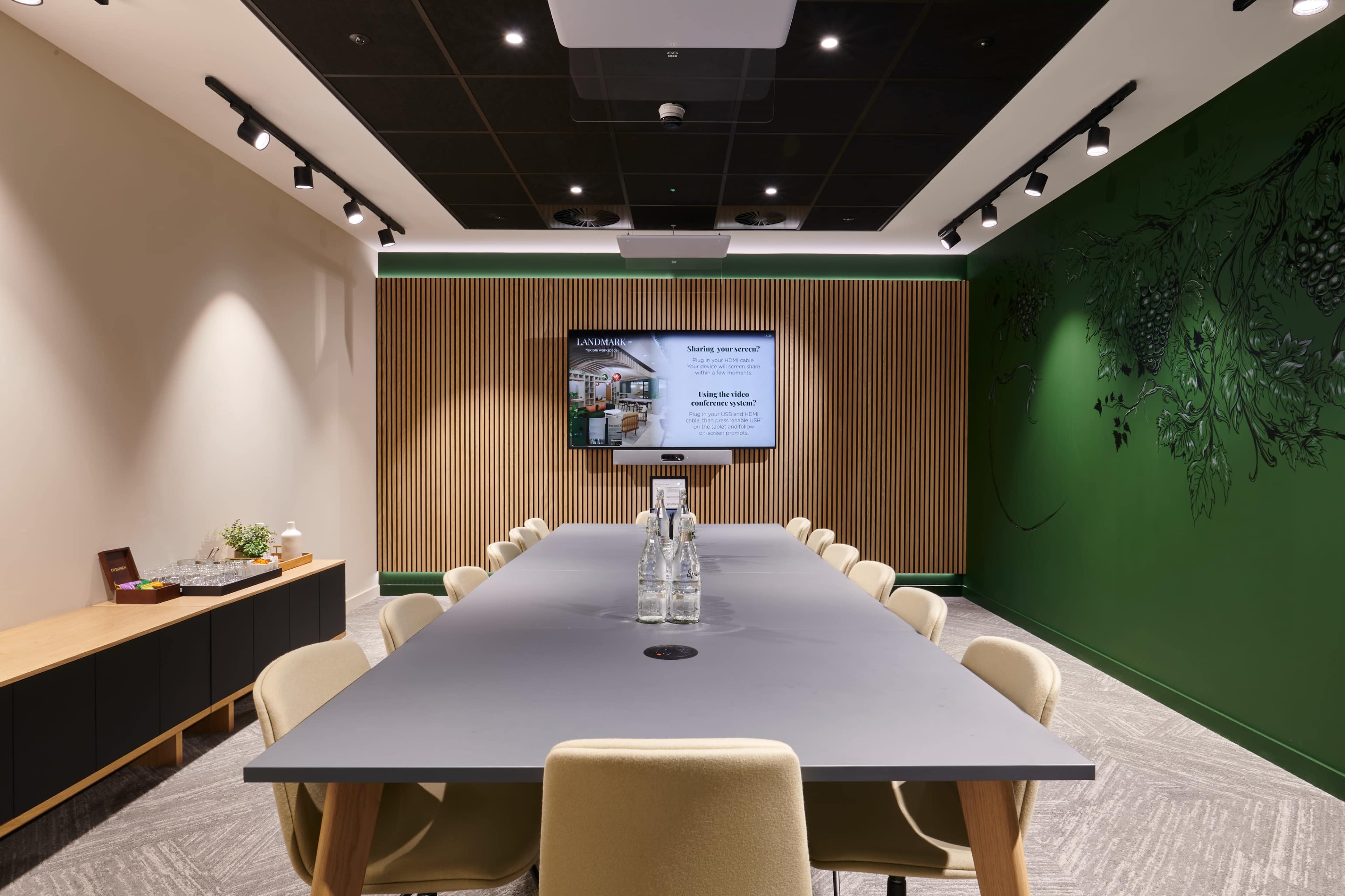 A long conference table is arranged in a modern meeting room with a screen mounted on the wall and green accent paint behind it.