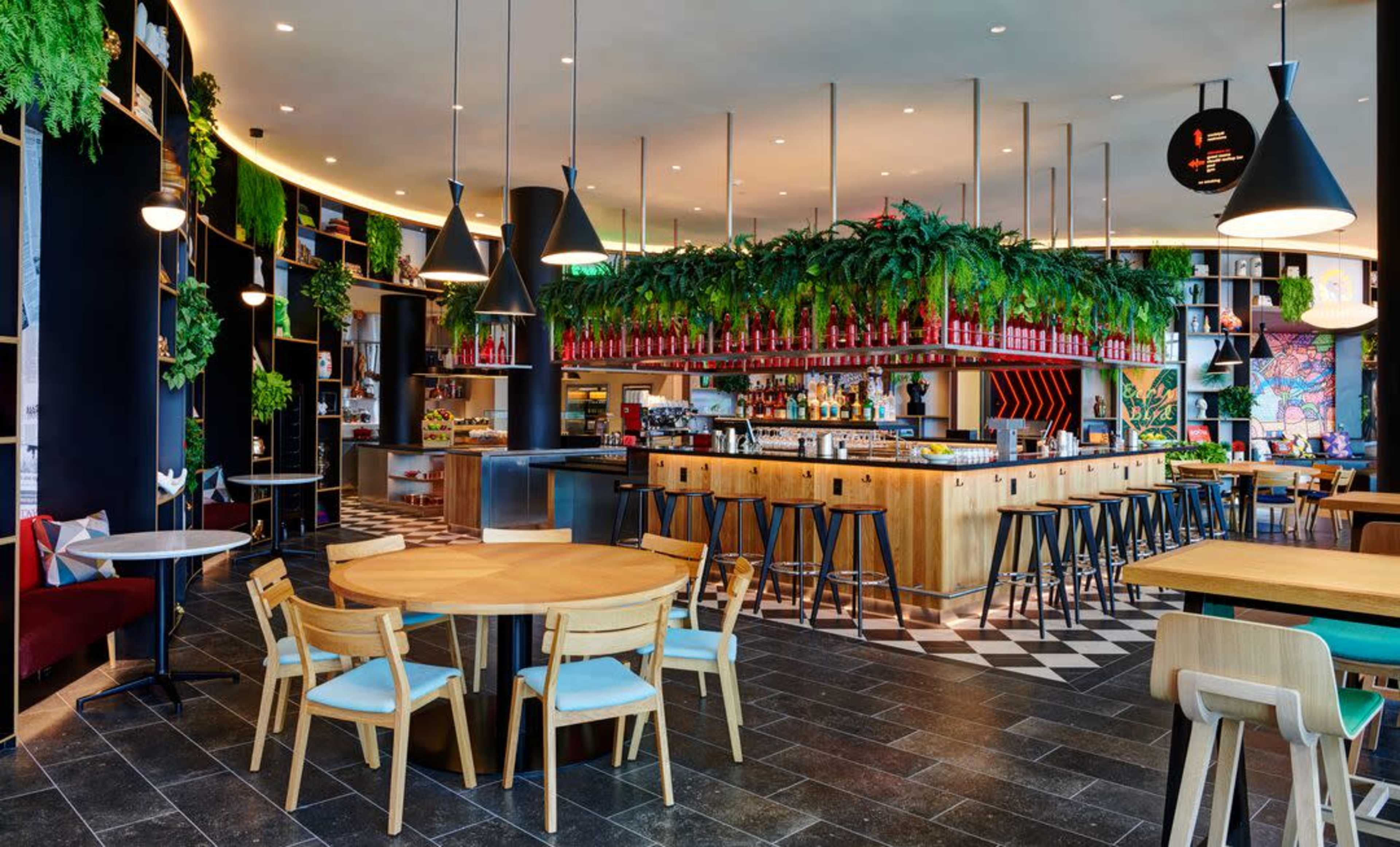 A modern restaurant interior featuring a stylish bar, wooden tables, and lush green plants hanging from the ceiling.