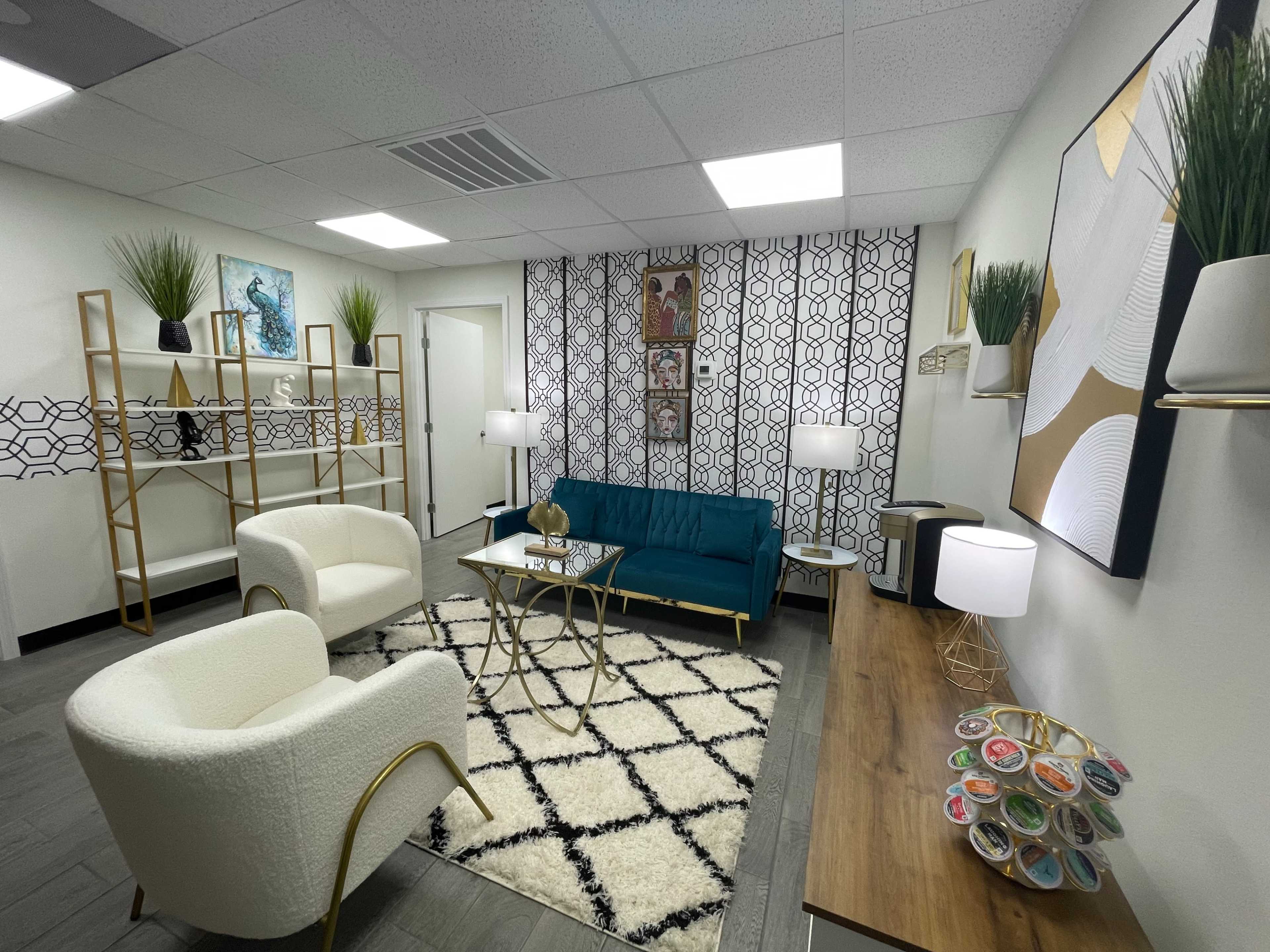 A modern waiting room featuring a teal sofa, two white armchairs, geometric wall decor, a coffee table, and decorative plants on shelves.