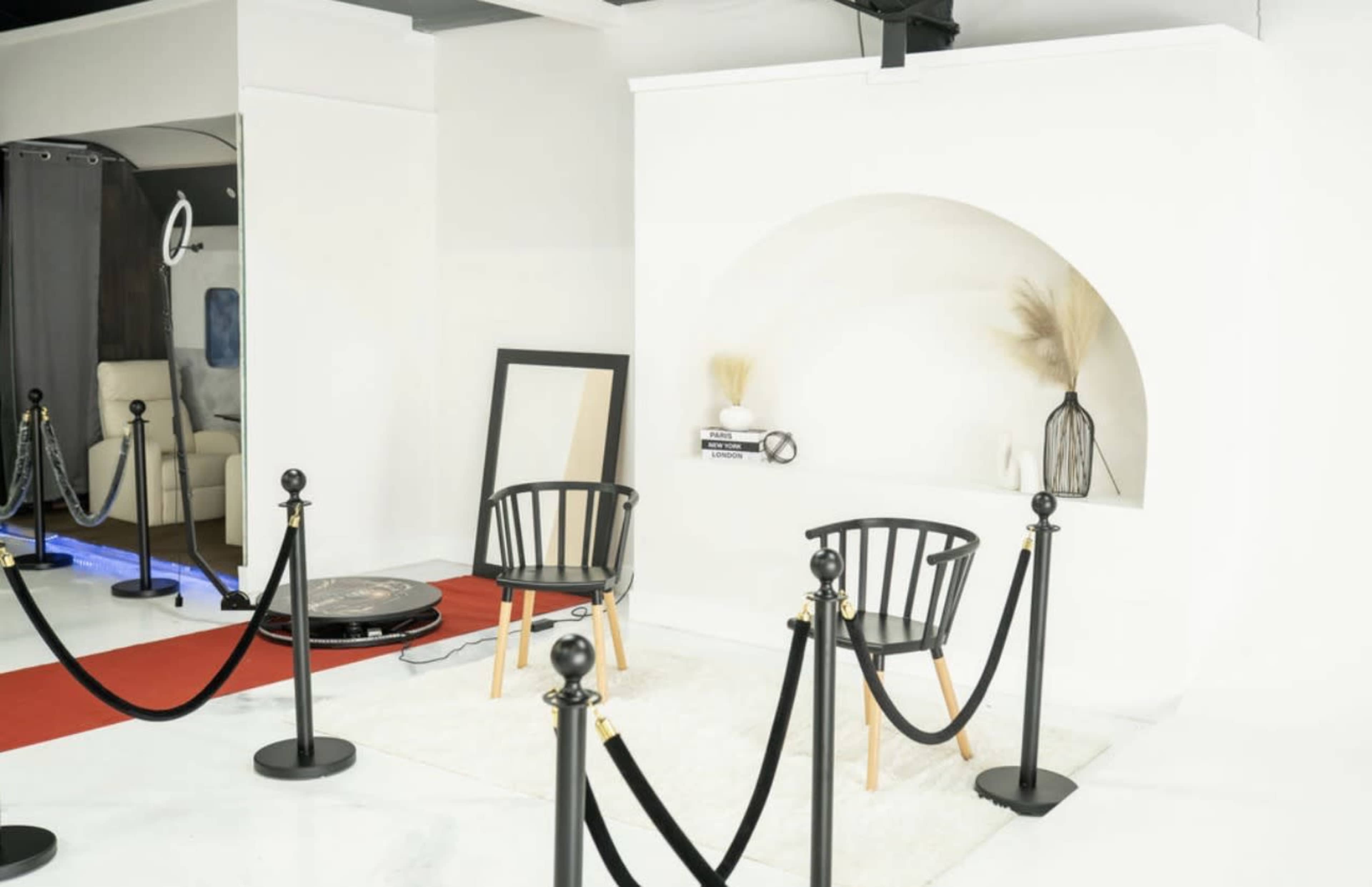 The image shows a minimalist interior with two black chairs on a white rug, a large mirror, and a decorative shelf with a vase and dried plant, all separated by a row of stanchions.