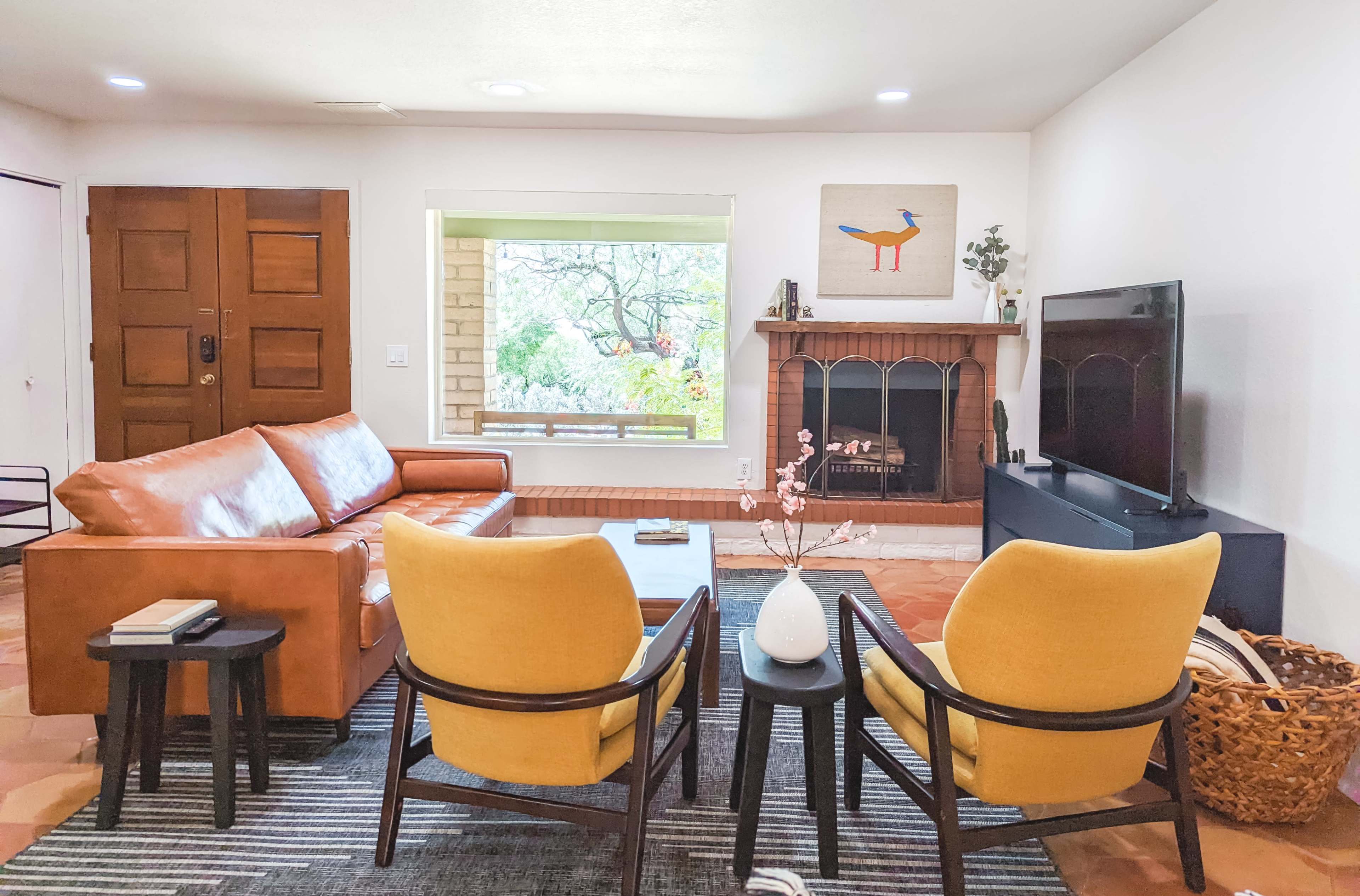 The living room features a brown leather sofa, two yellow chairs, a coffee table, and a television facing a decorative fireplace beneath a window.