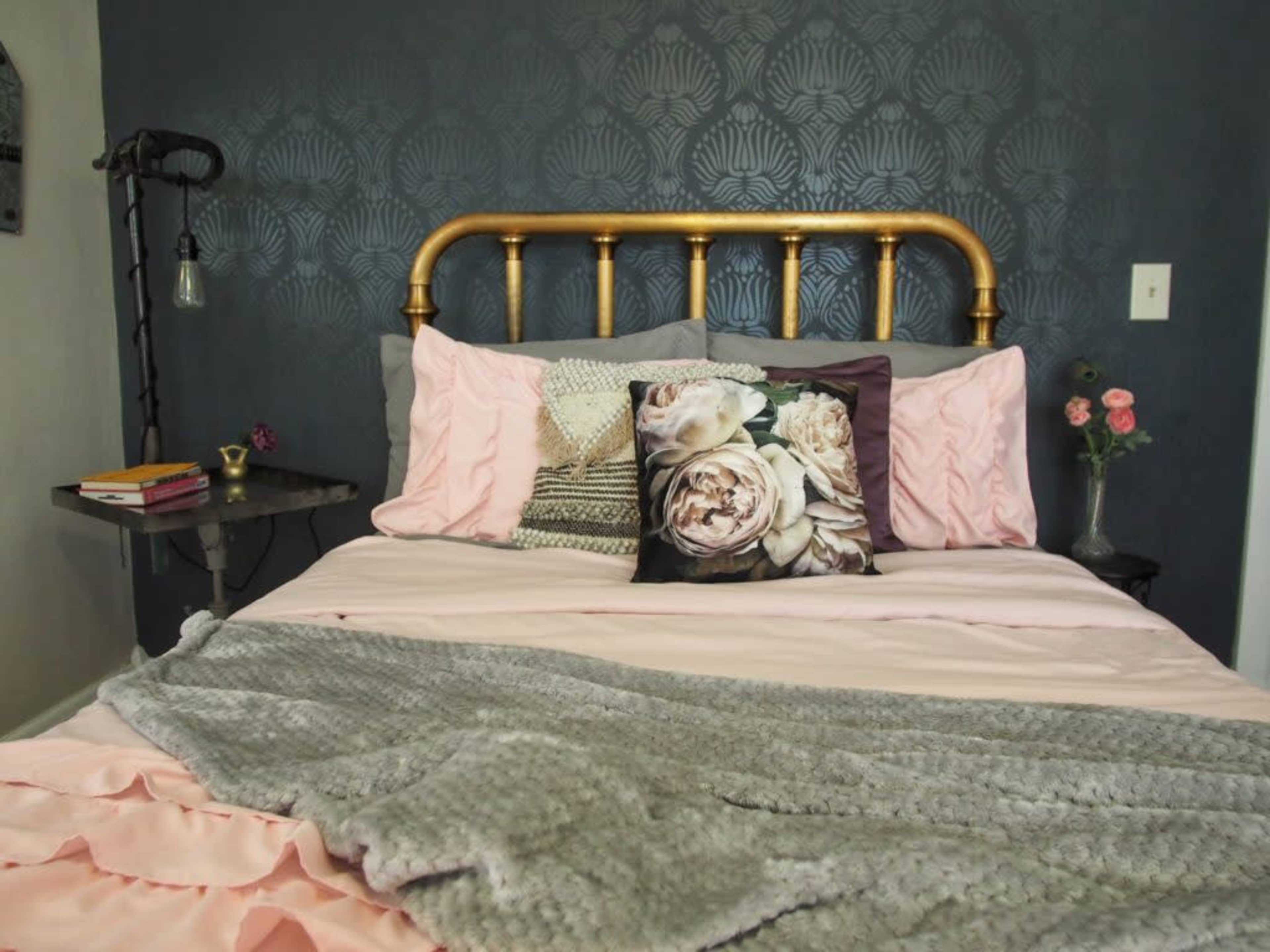 A neatly made bed features a gold metal headboard, decorative pillows, and a soft gray blanket against a dark patterned wall.