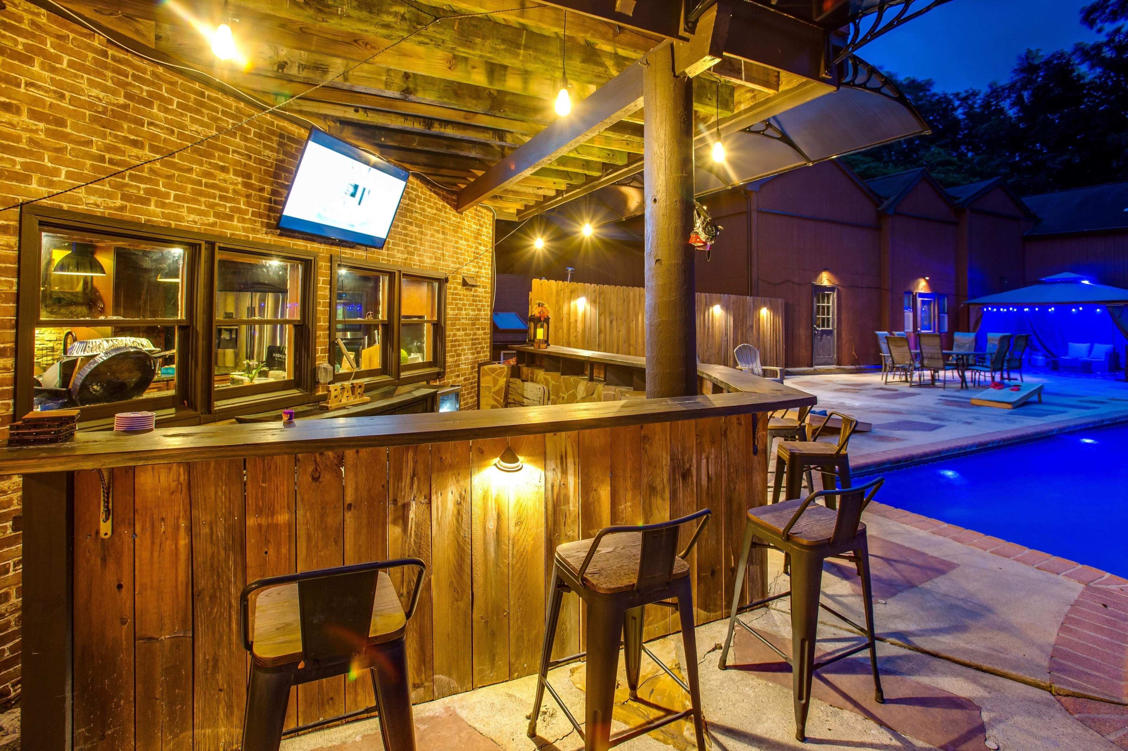 A dimly lit outdoor bar area with wooden countertops and stools, overlooking a swimming pool surrounded by lounge chairs and a nearby building.