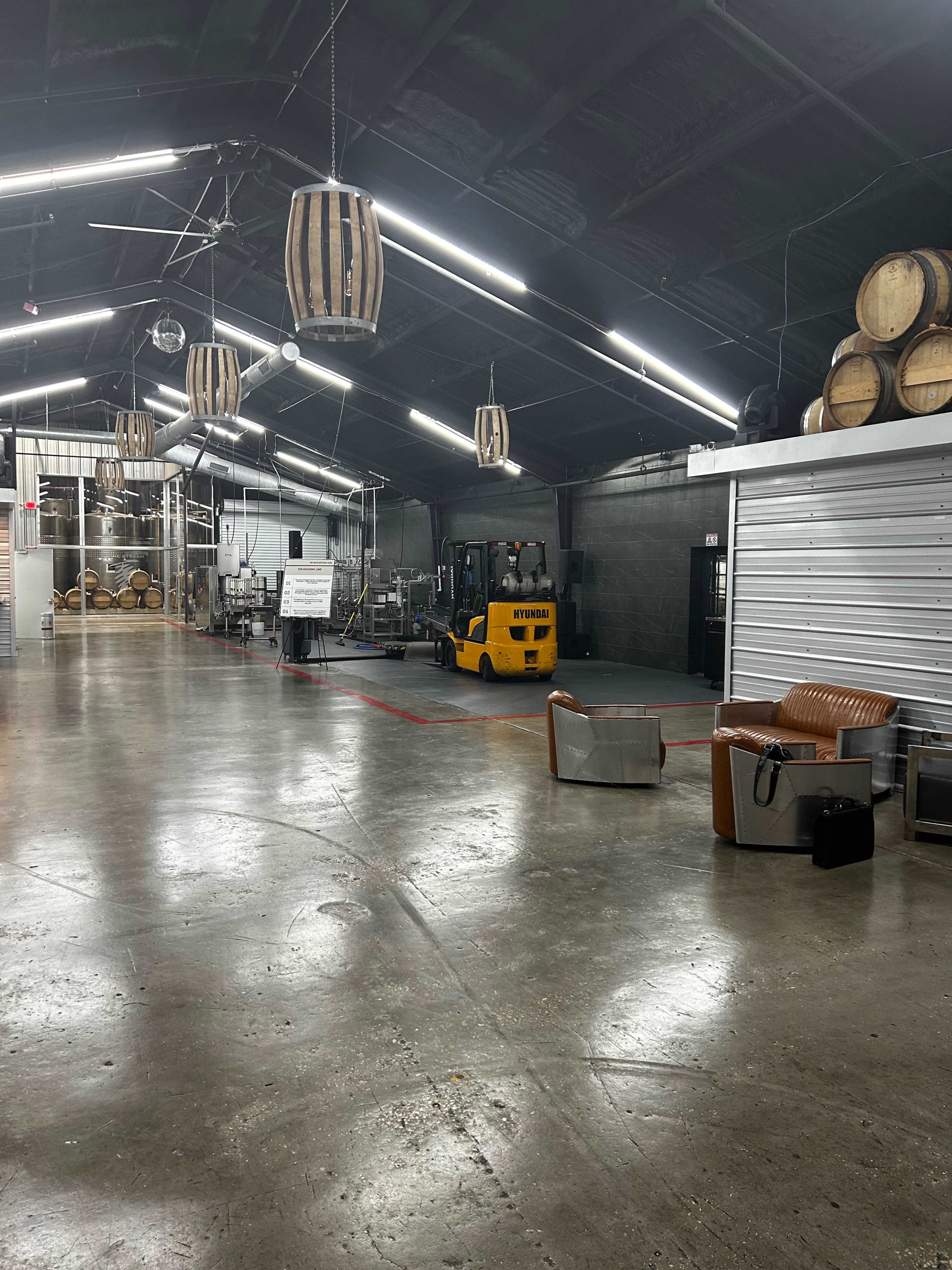 The image shows an industrial space with wooden barrels, a forklift, and various seating arrangements, illuminated by overhead lights.