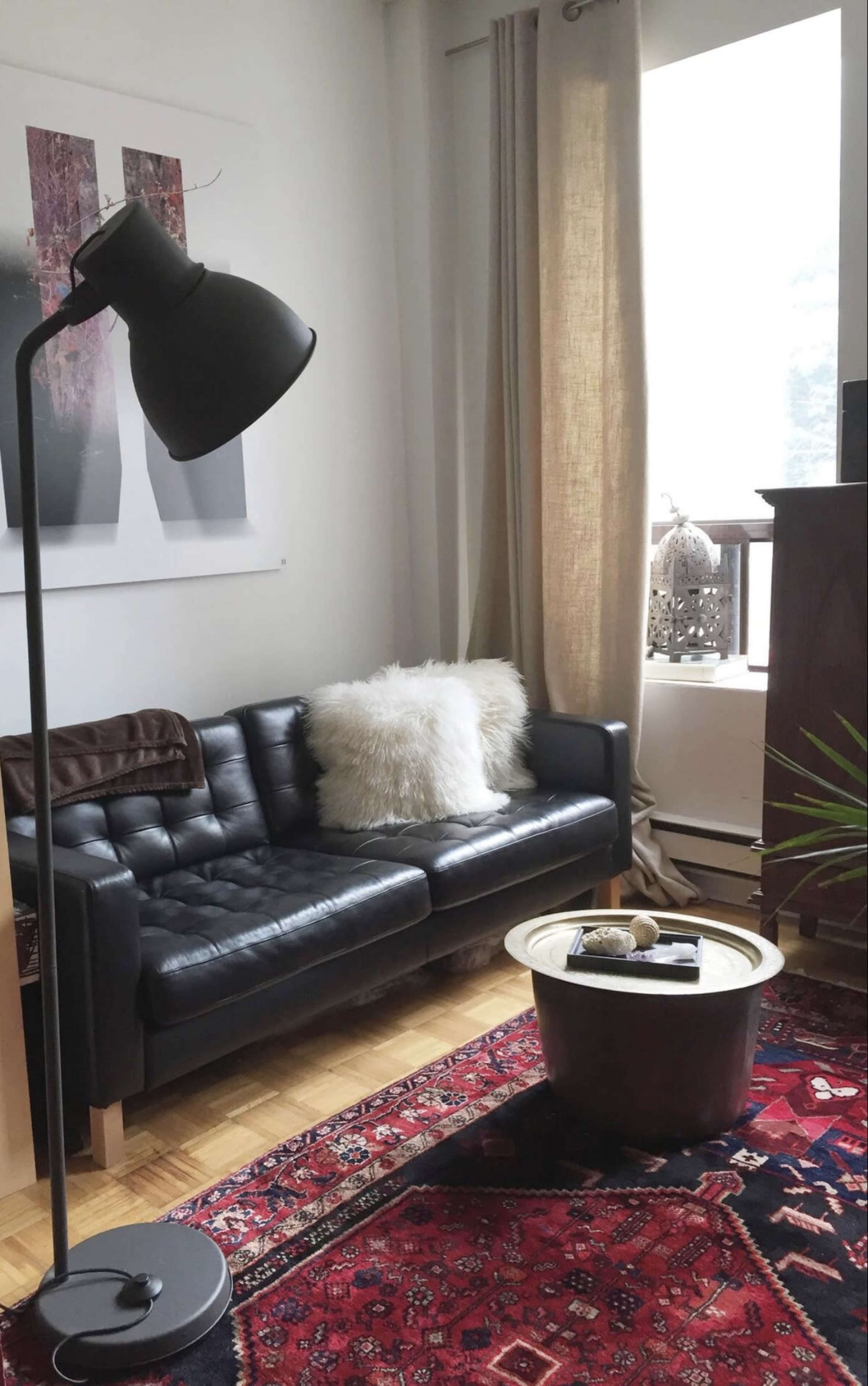 A cozy living room with a black leather couch, a plush white throw pillow, a round coffee table, and a floor lamp, all set on a patterned rug.