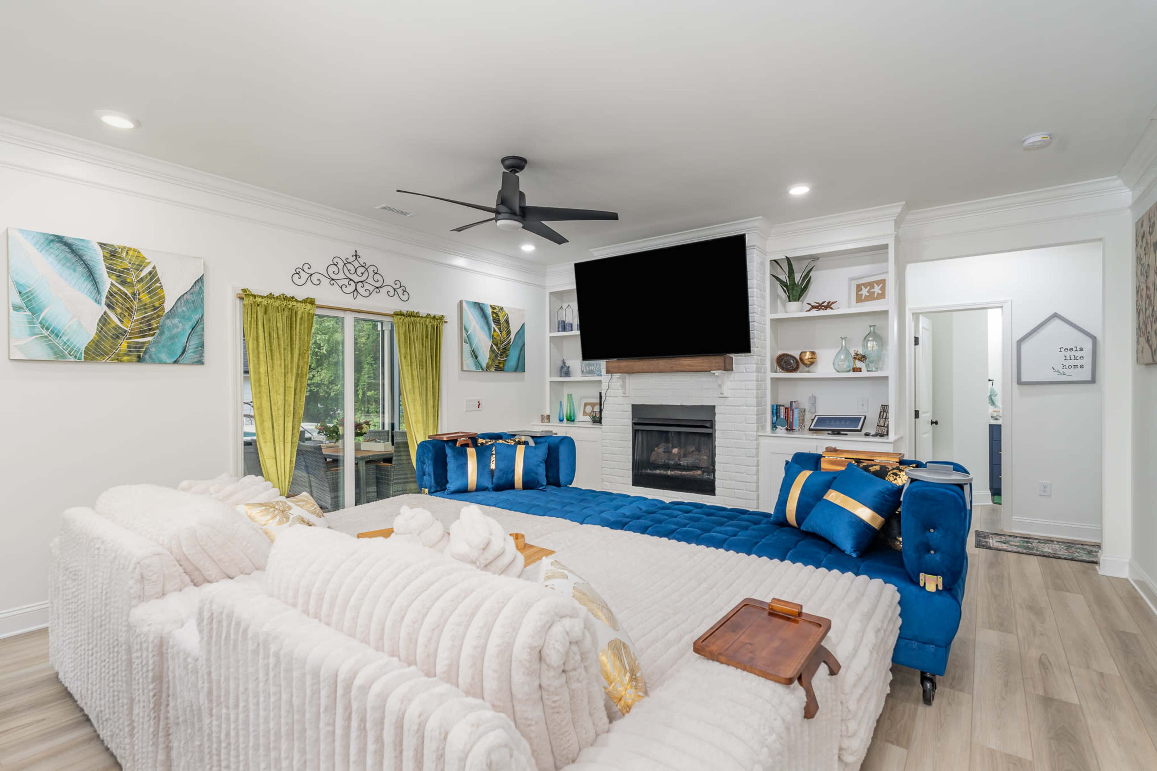A modern living room featuring a blue tufted sofa, a fireplace with a white mantel, open shelving, and large windows with green curtains.