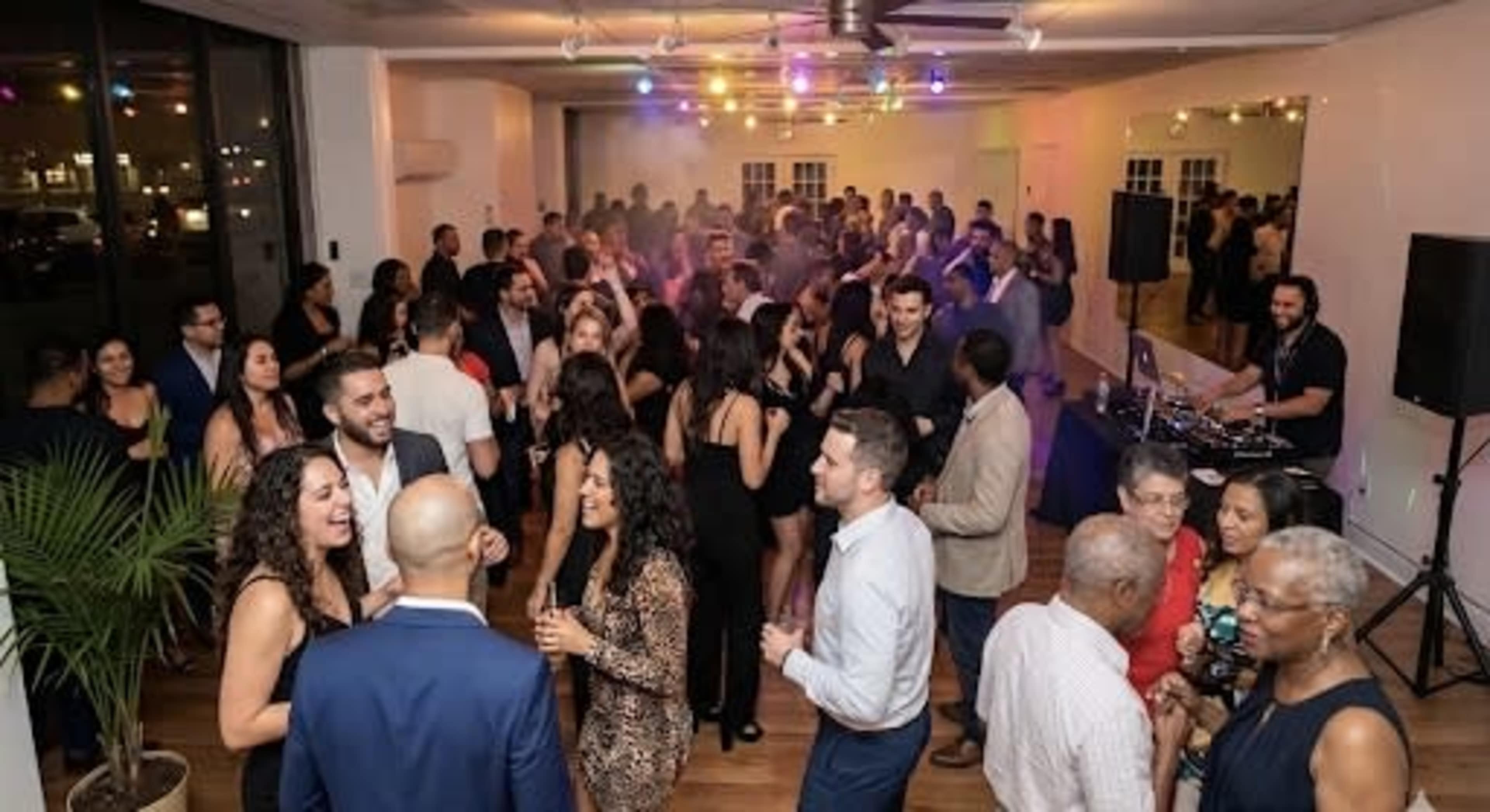 A lively crowd of people dances in a well-lit room with festive lighting and a DJ setup in the background.