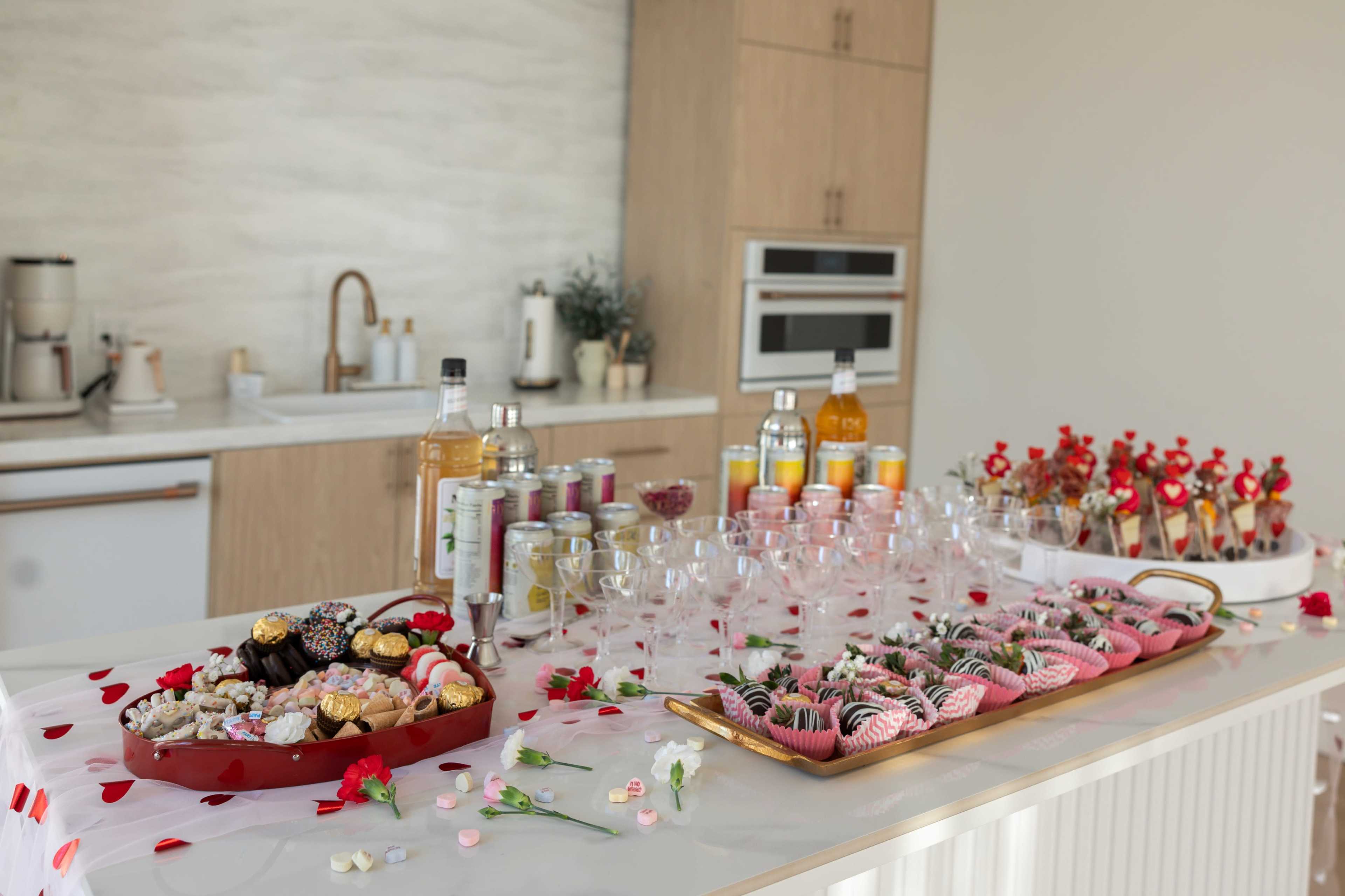 A buffet-style table is set with an assortment of sweets, drinks, and decorative rose petals in a modern kitchen.