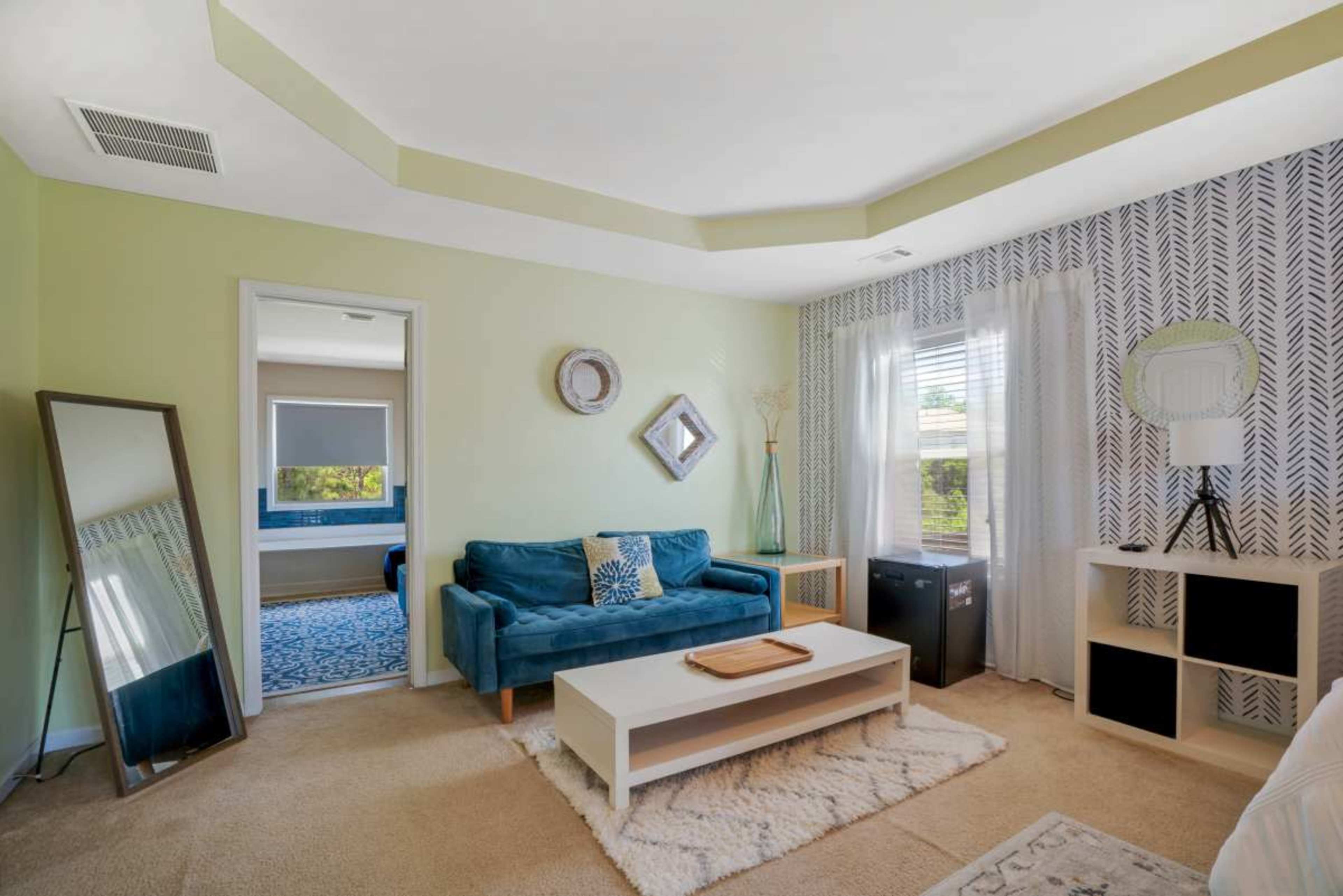 A small living space features a teal sofa, a coffee table, a mirror, and shelving against a light green wall with patterned wallpaper.