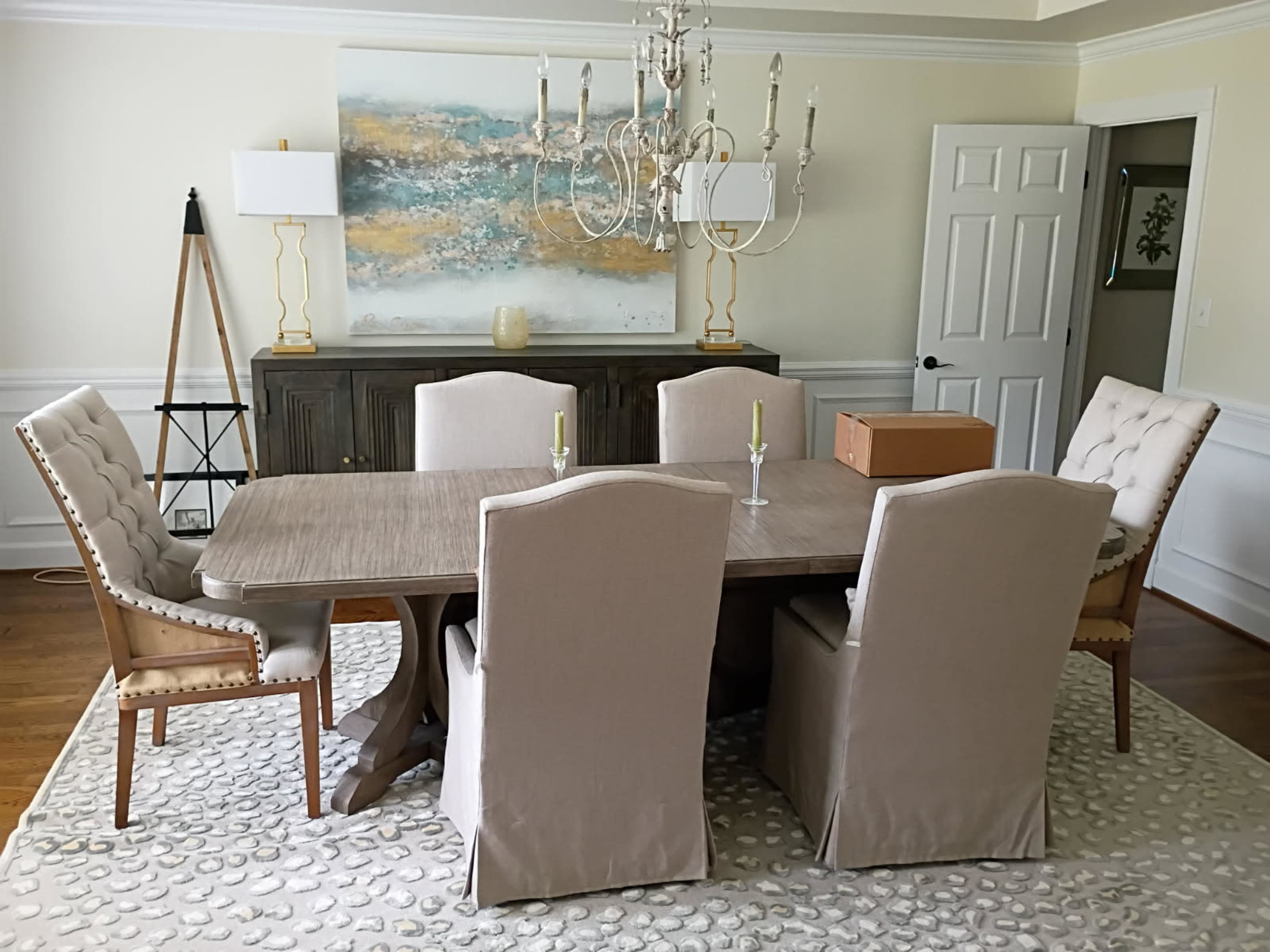 A modern dining room features a large wooden table surrounded by six upholstered chairs, a chandelier overhead, and a sideboard against the wall.