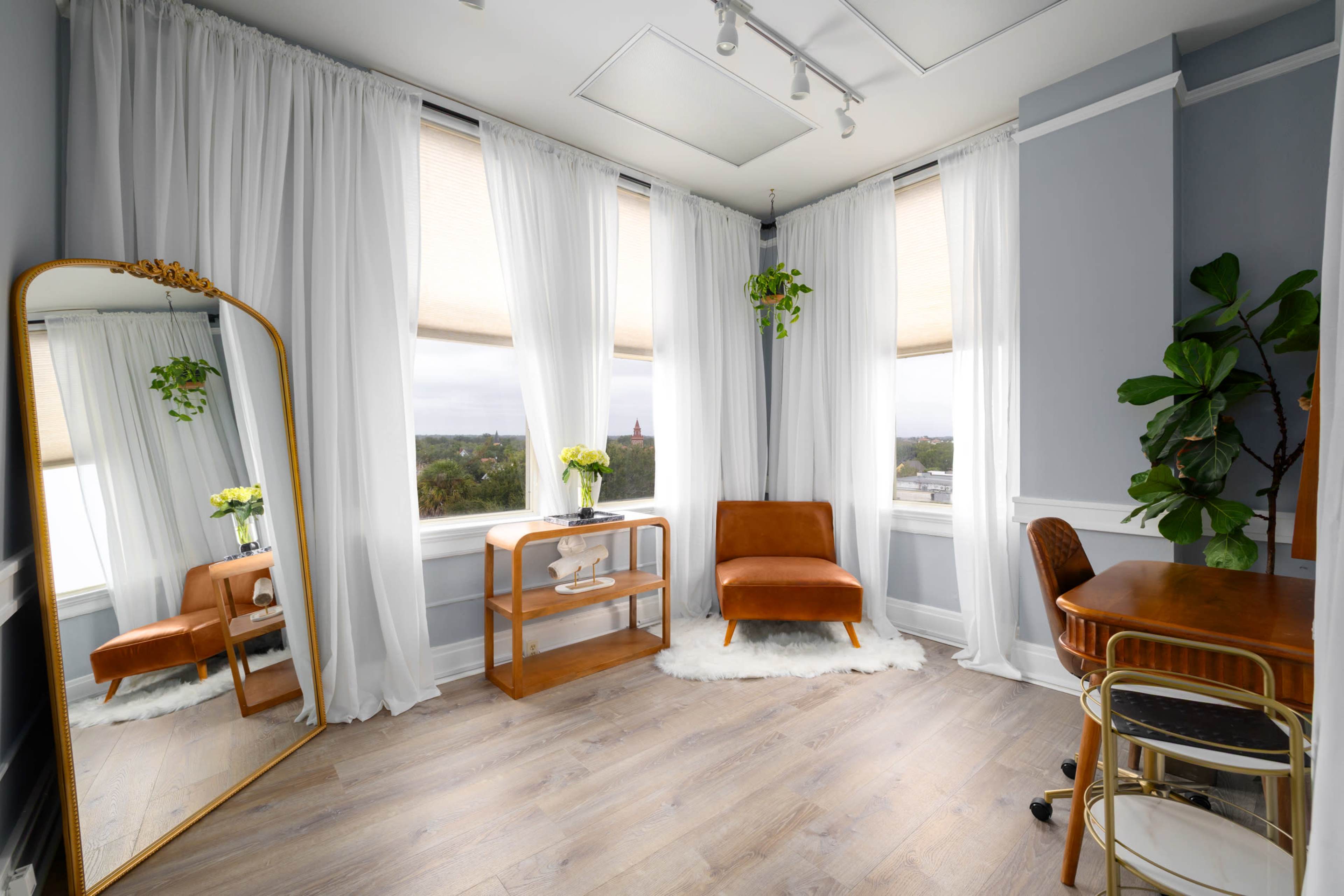 The image shows a well-lit room featuring a brown accent chair, a wooden table, a large mirror, and potted plants, with sheer white curtains covering the windows.