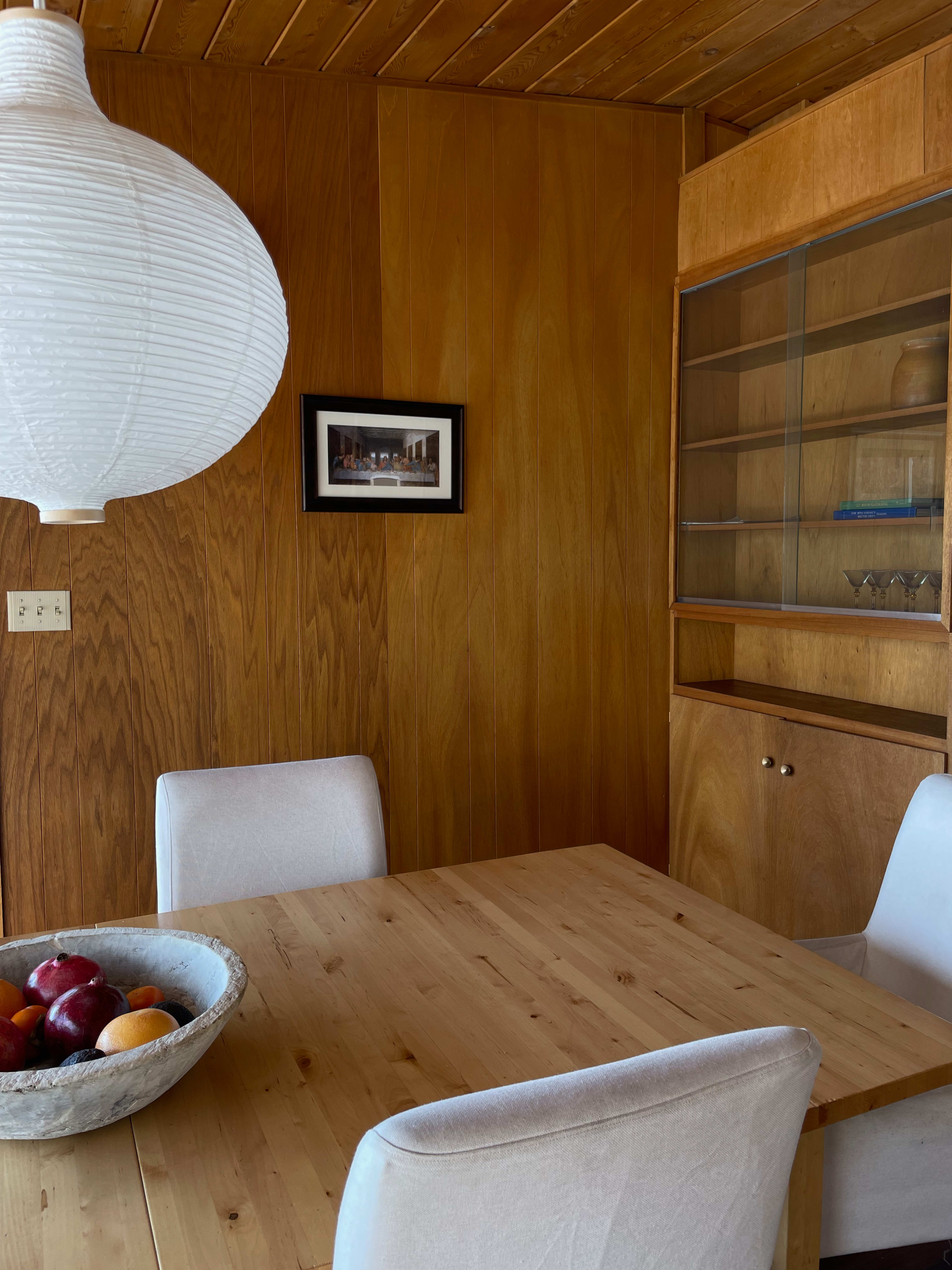 A dining area with a wooden table, white chairs, a bowl of fruit, a paper lantern, and a wooden paneled wall with a framed photograph.