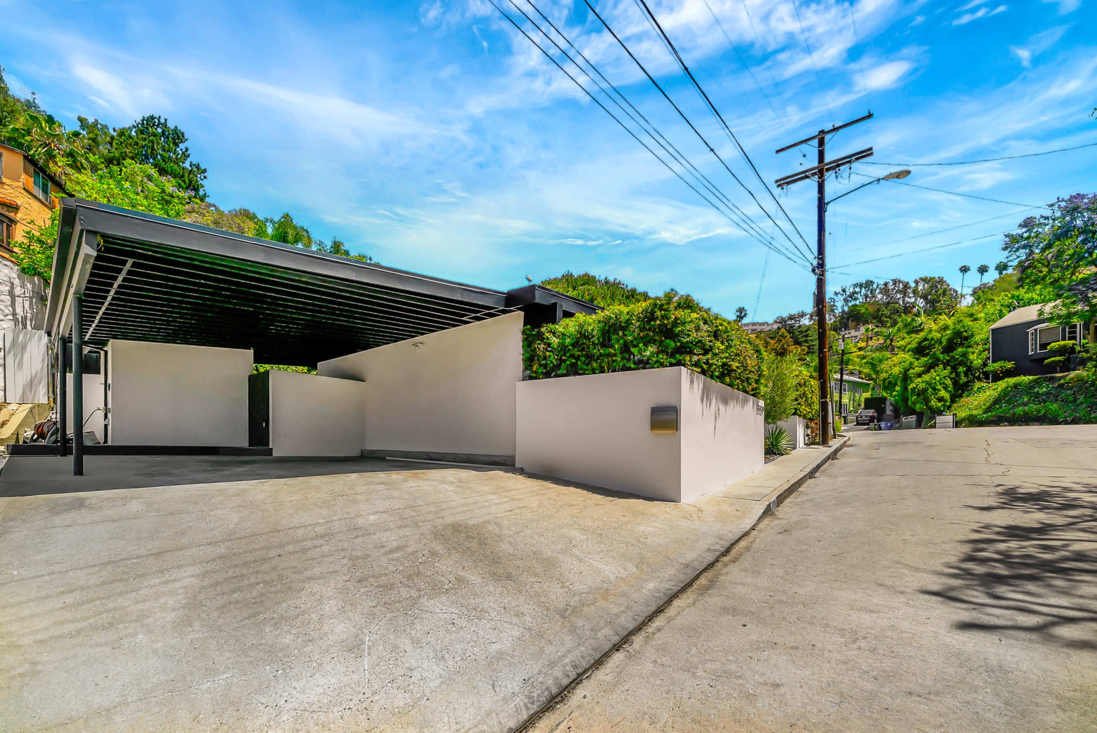 A modern carport with a white concrete wall is situated alongside a residential street, surrounded by greenery and power lines.