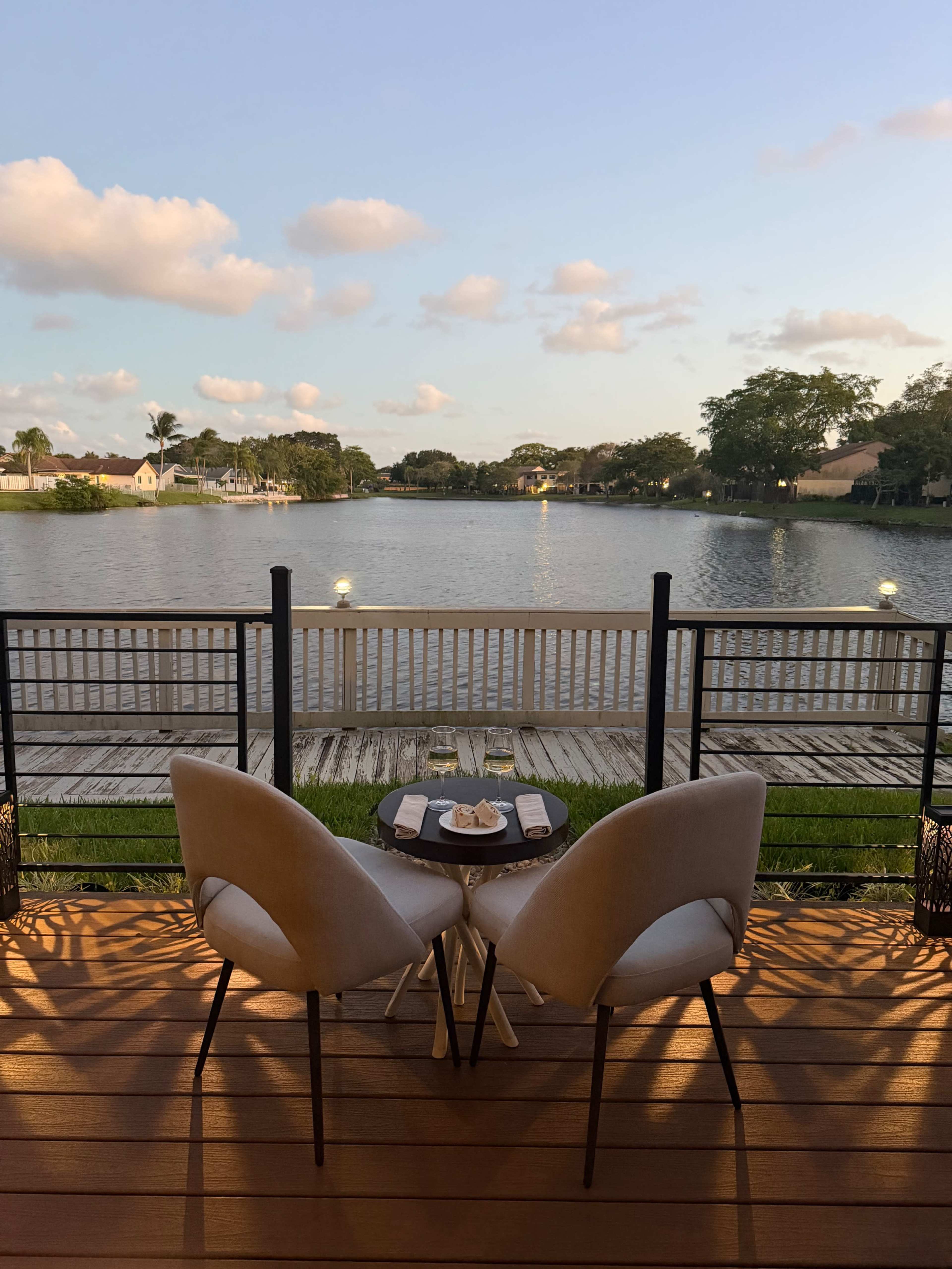 A small table set for two with white chairs overlooks a calm lake, framed by a wooden deck and gently swaying trees in the background.