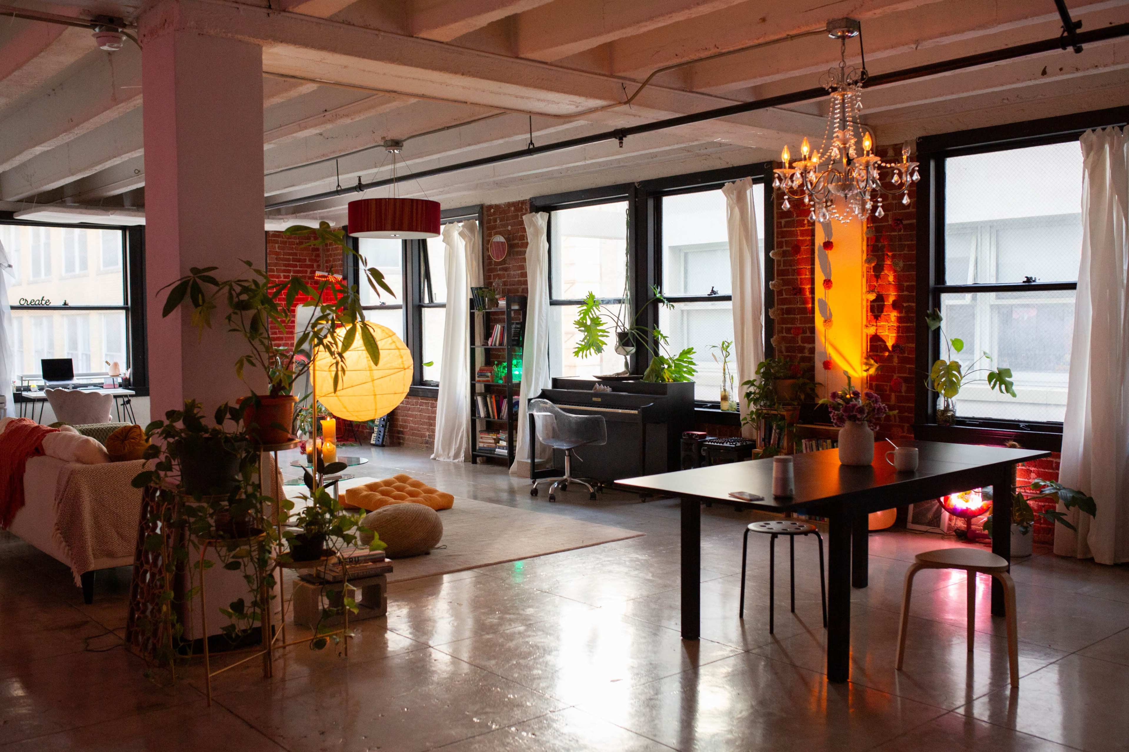 The image shows a spacious, modern living area with large windows, a dining table, various indoor plants, and a cozy seating arrangement.