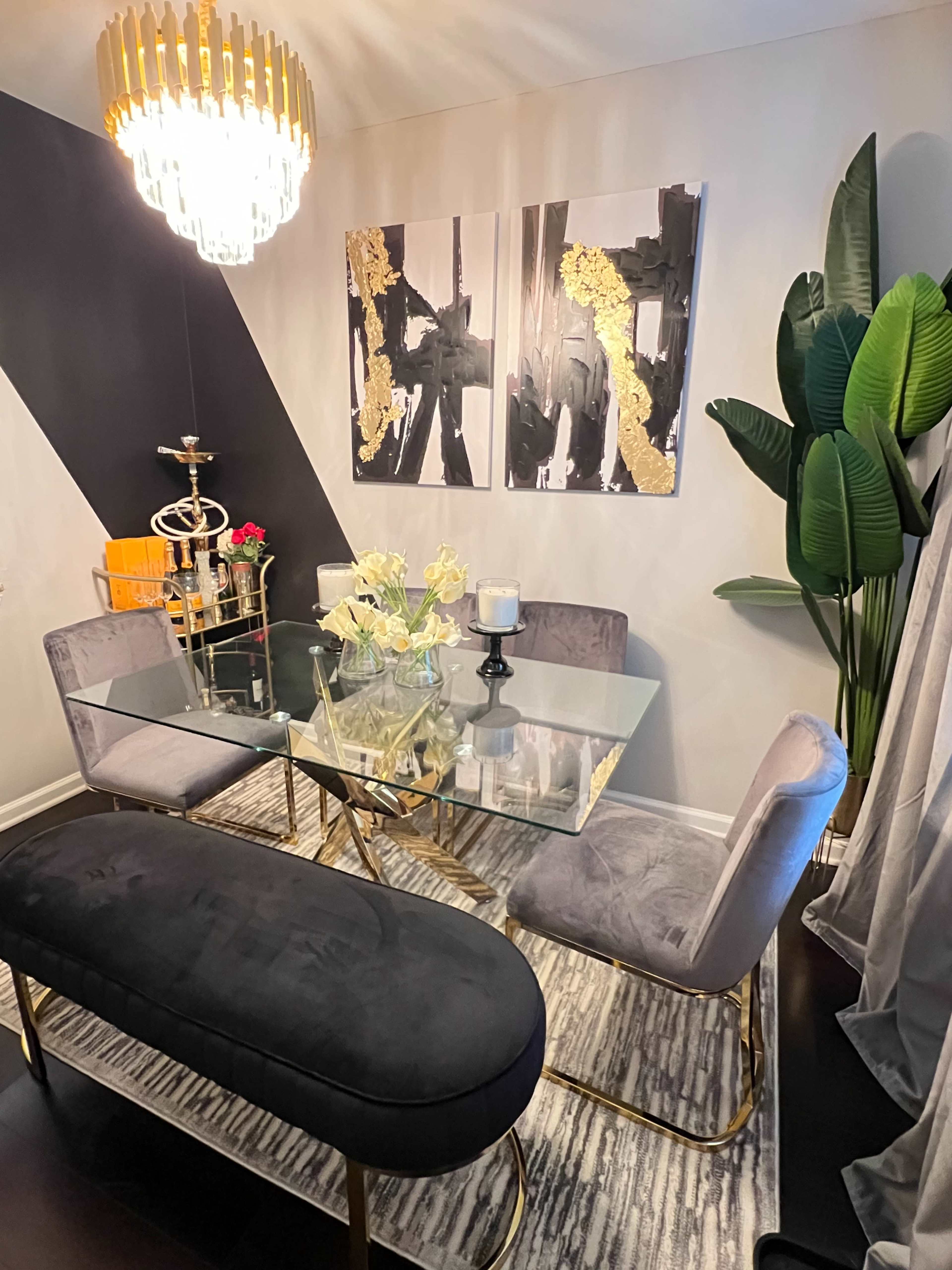 A modern dining area features a glass table surrounded by upholstered chairs, a bench, and wall art, illuminated by a chandelier.