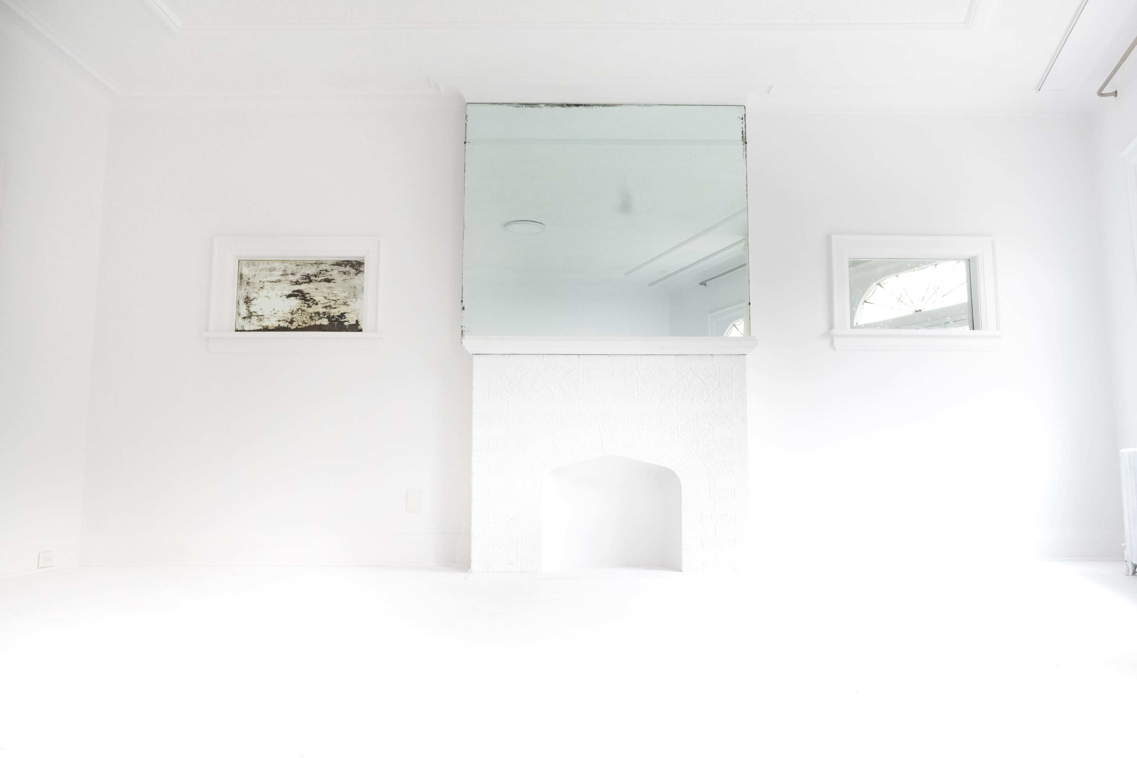 A minimalistic white room features a large mirror above a white fireplace, with two small windows on either side.