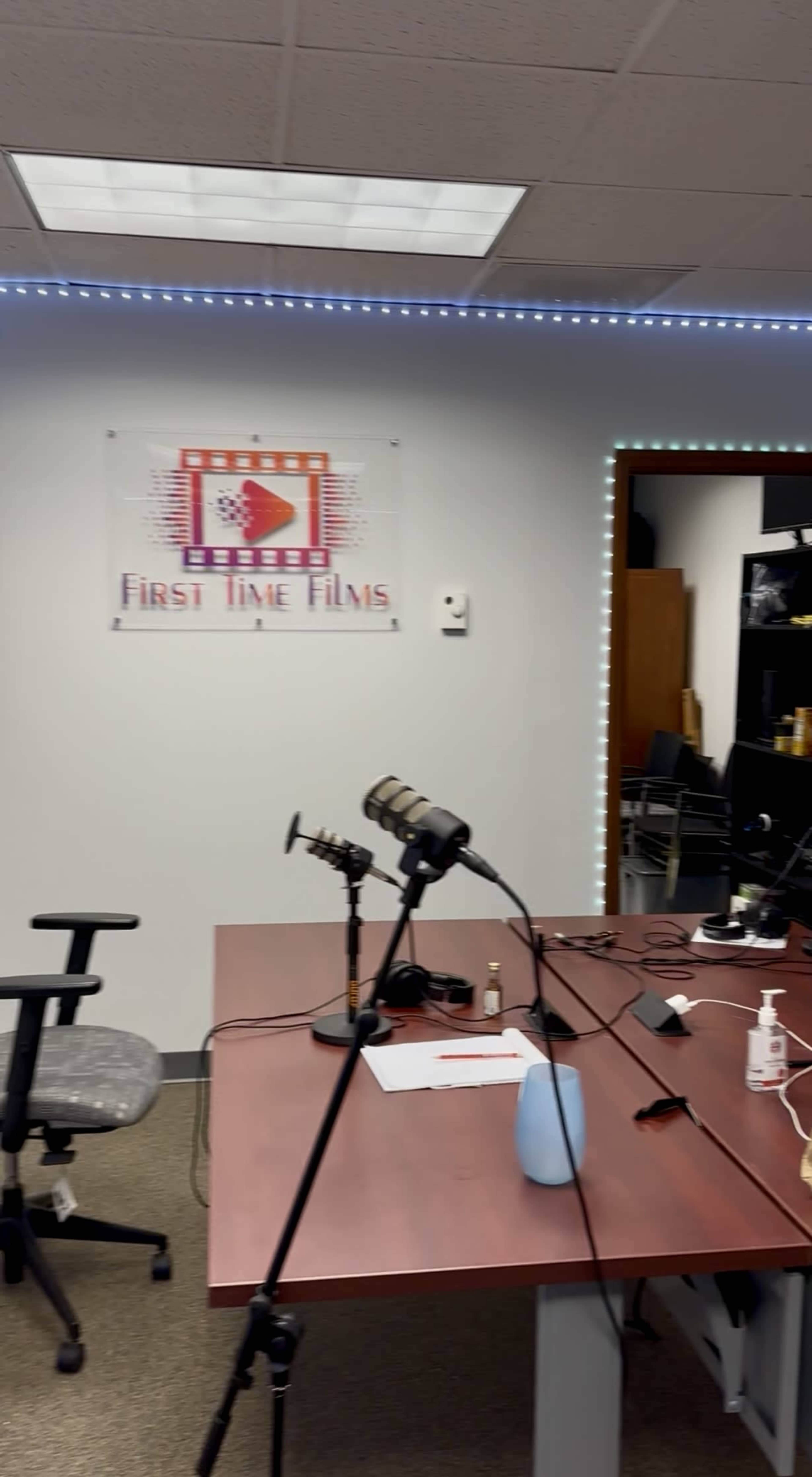 The image shows a film studio or podcast recording space with a table, microphones, and a sign for "First Time Films" on the wall.