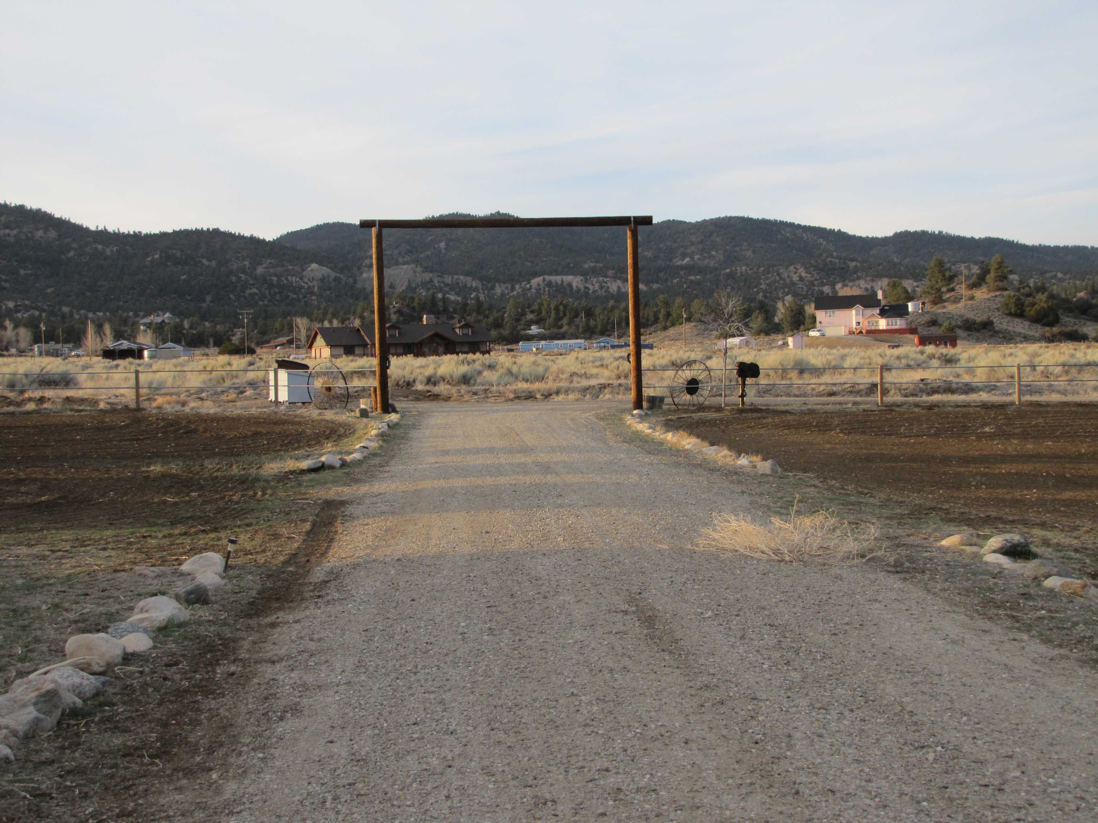 A gravel road leads to a wooden archway at the entrance of a rural property, with houses visible in the background and mountains in the distance.