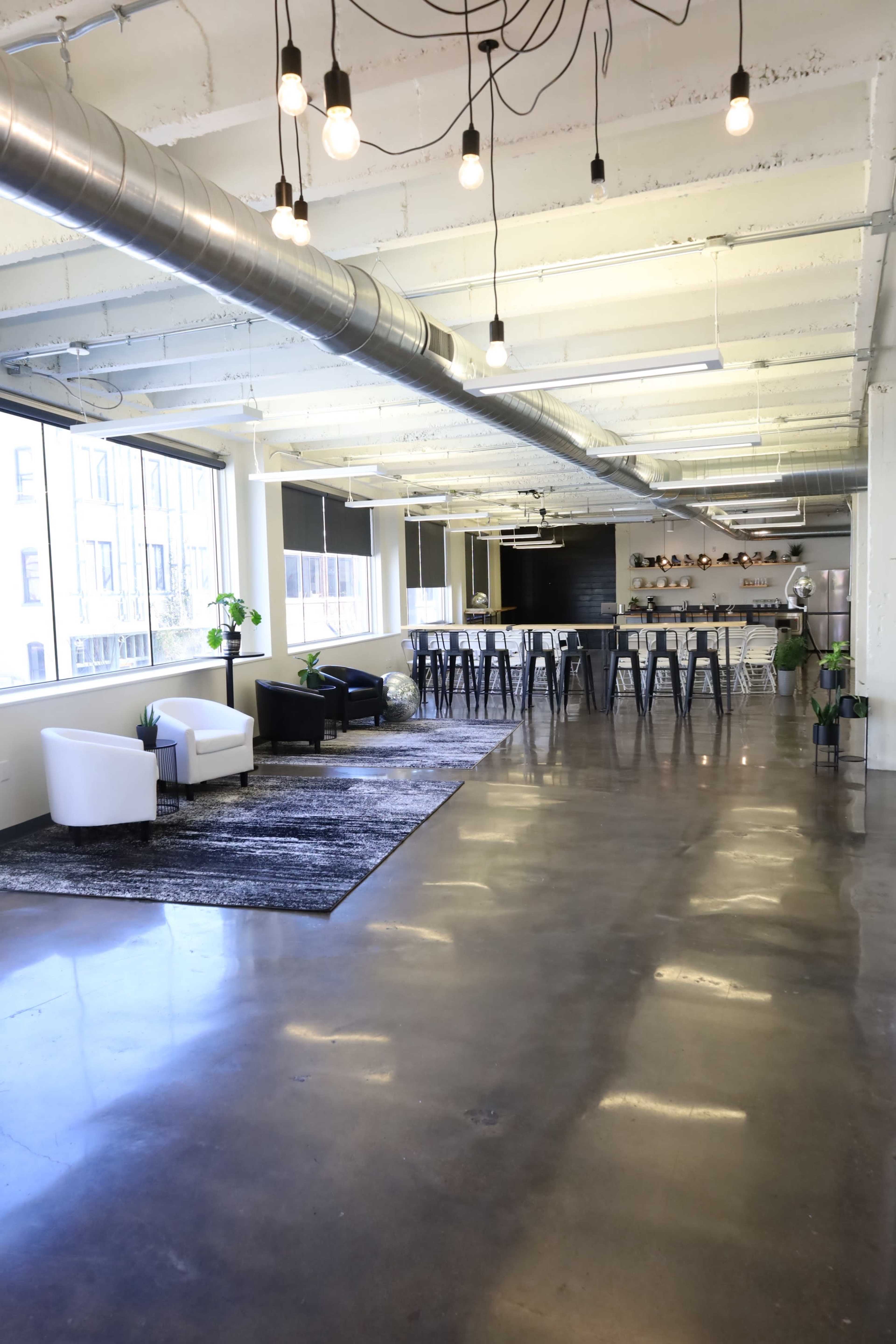 A spacious, modern office interior with polished concrete floors, a seating area with two armchairs, and a long bar-style table with metal stools.