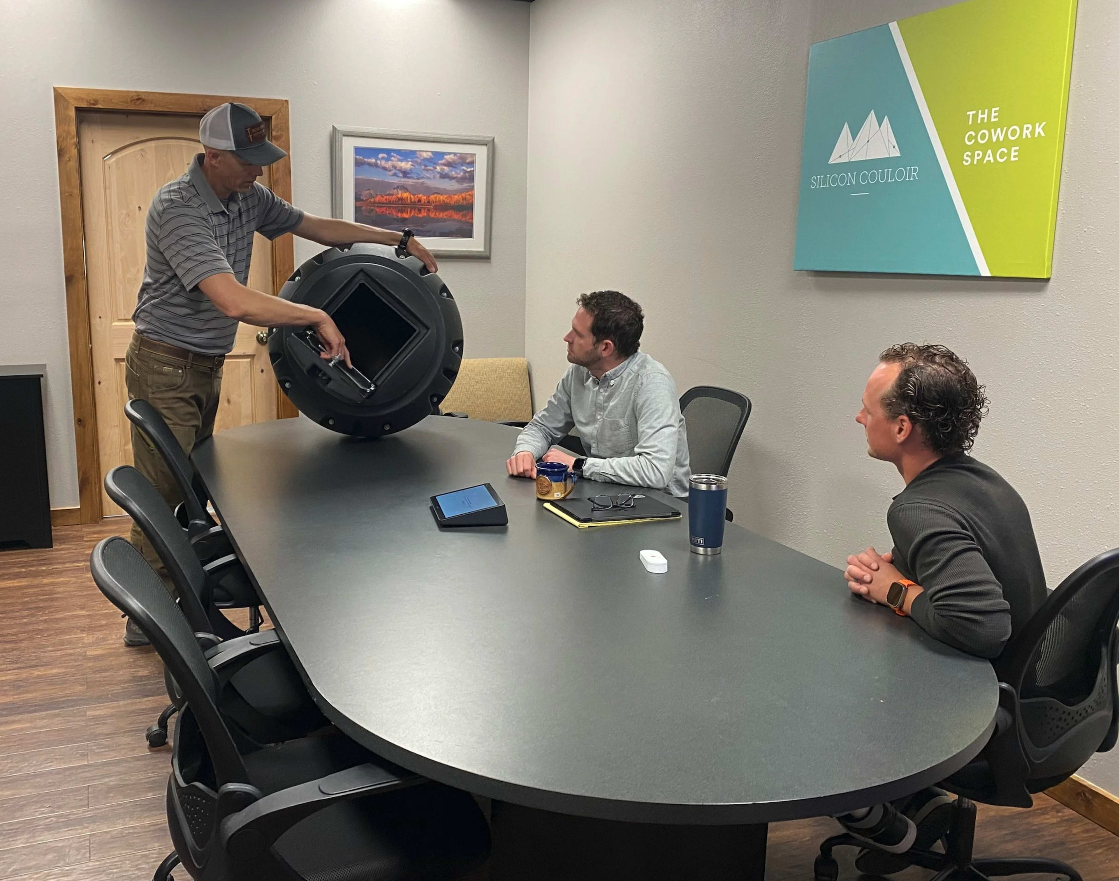 A person is demonstrating a device to two seated colleagues at a table in a conference room.