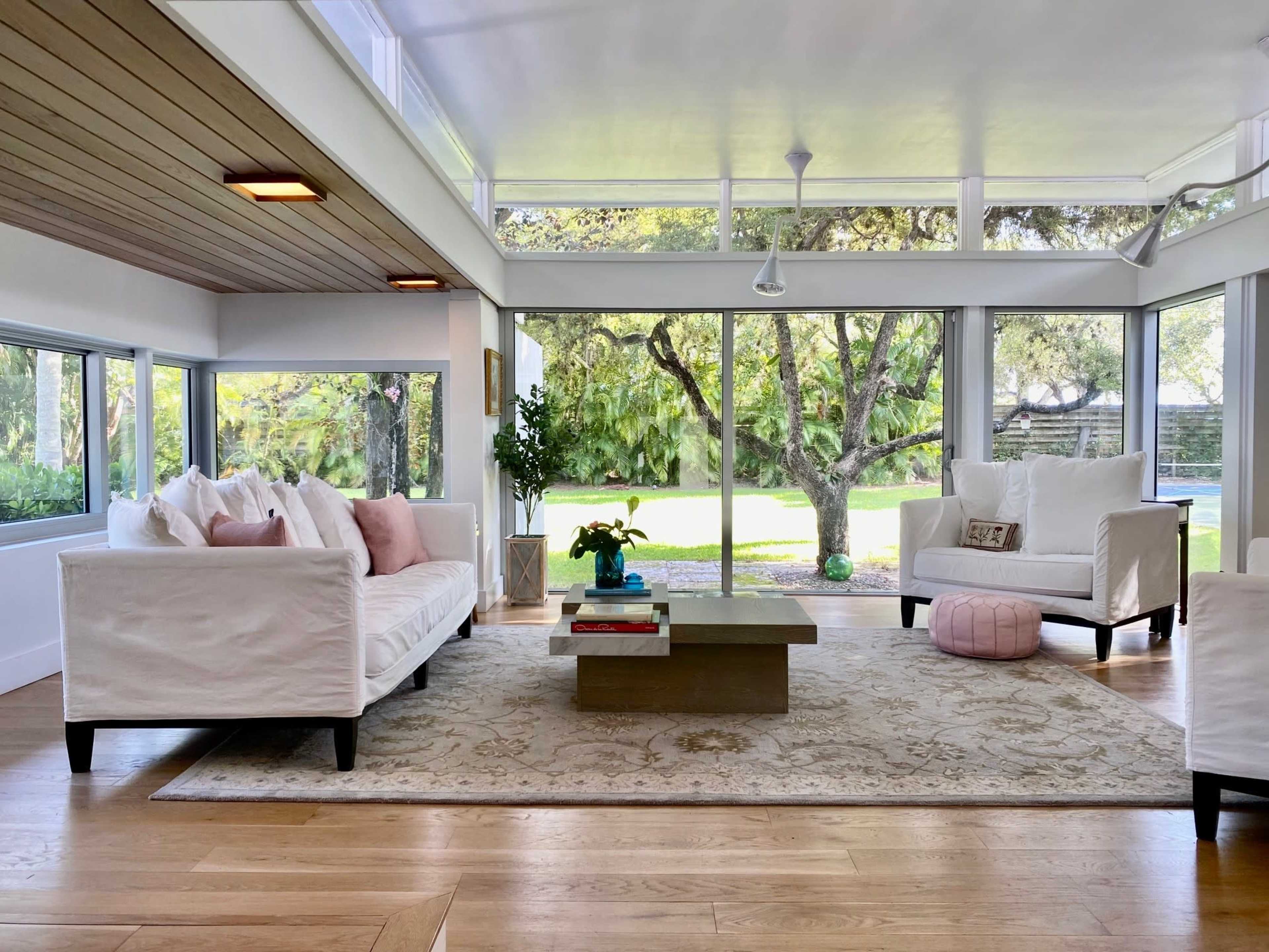 A bright, airy living room features large windows, a comfortable seating arrangement, and a central coffee table on a decorative rug.