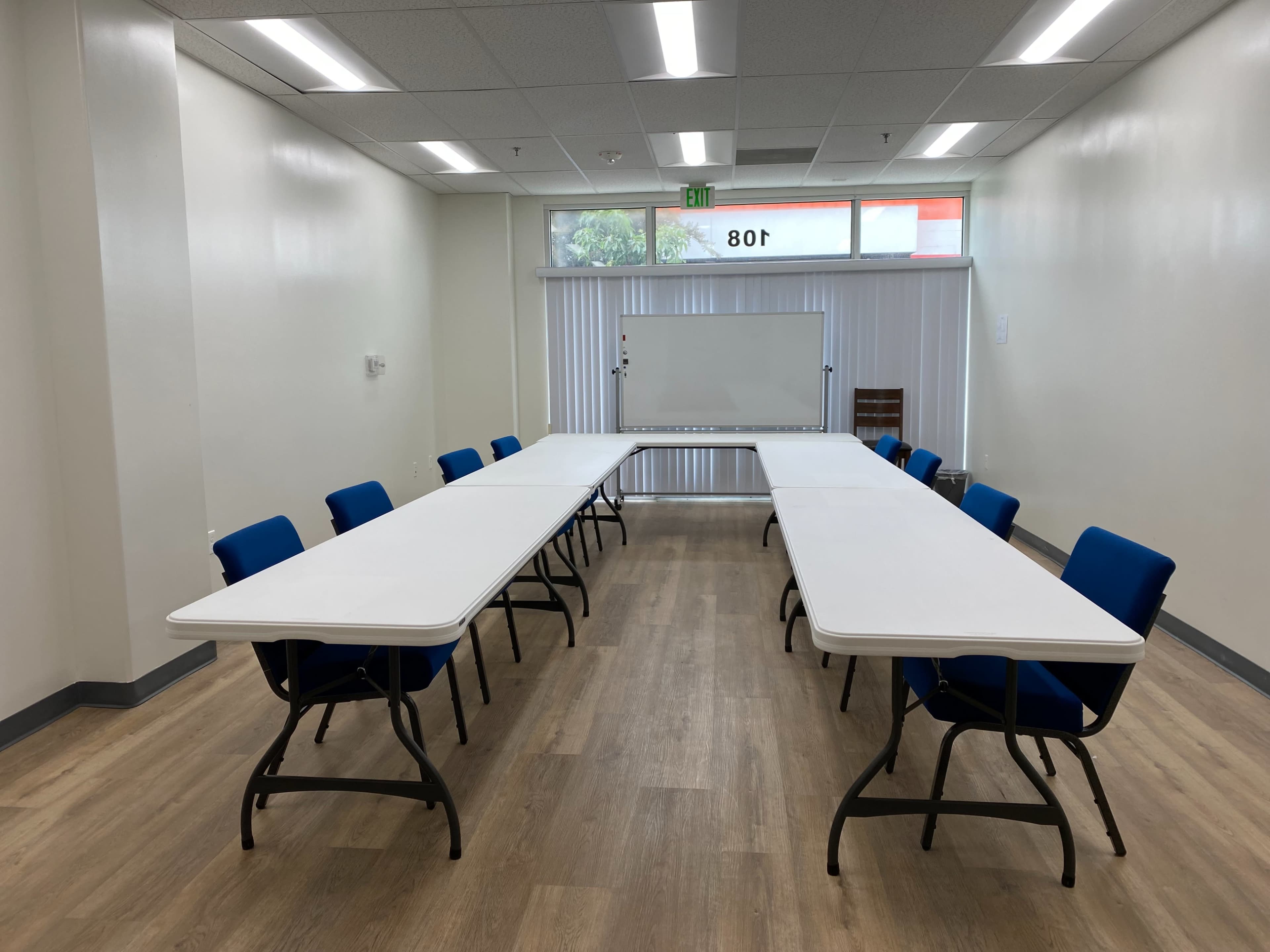 A training room features two sets of rectangular tables arranged in a U-shape, surrounded by blue chairs, with large windows allowing natural light.