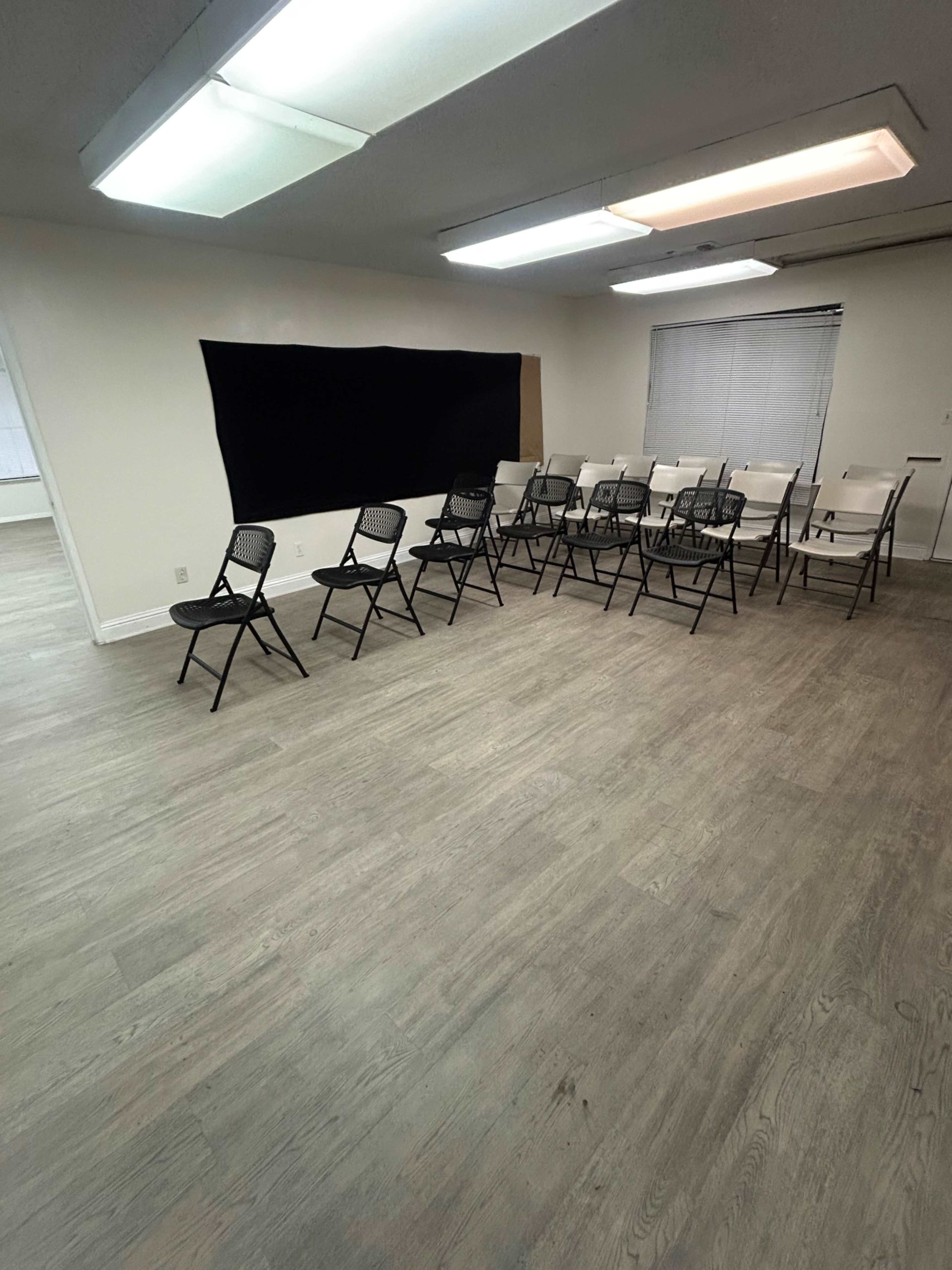 The image shows a room with a row of eleven folding chairs arranged in front of a black backdrop, with large windows on one side and fluorescent ceiling lights.