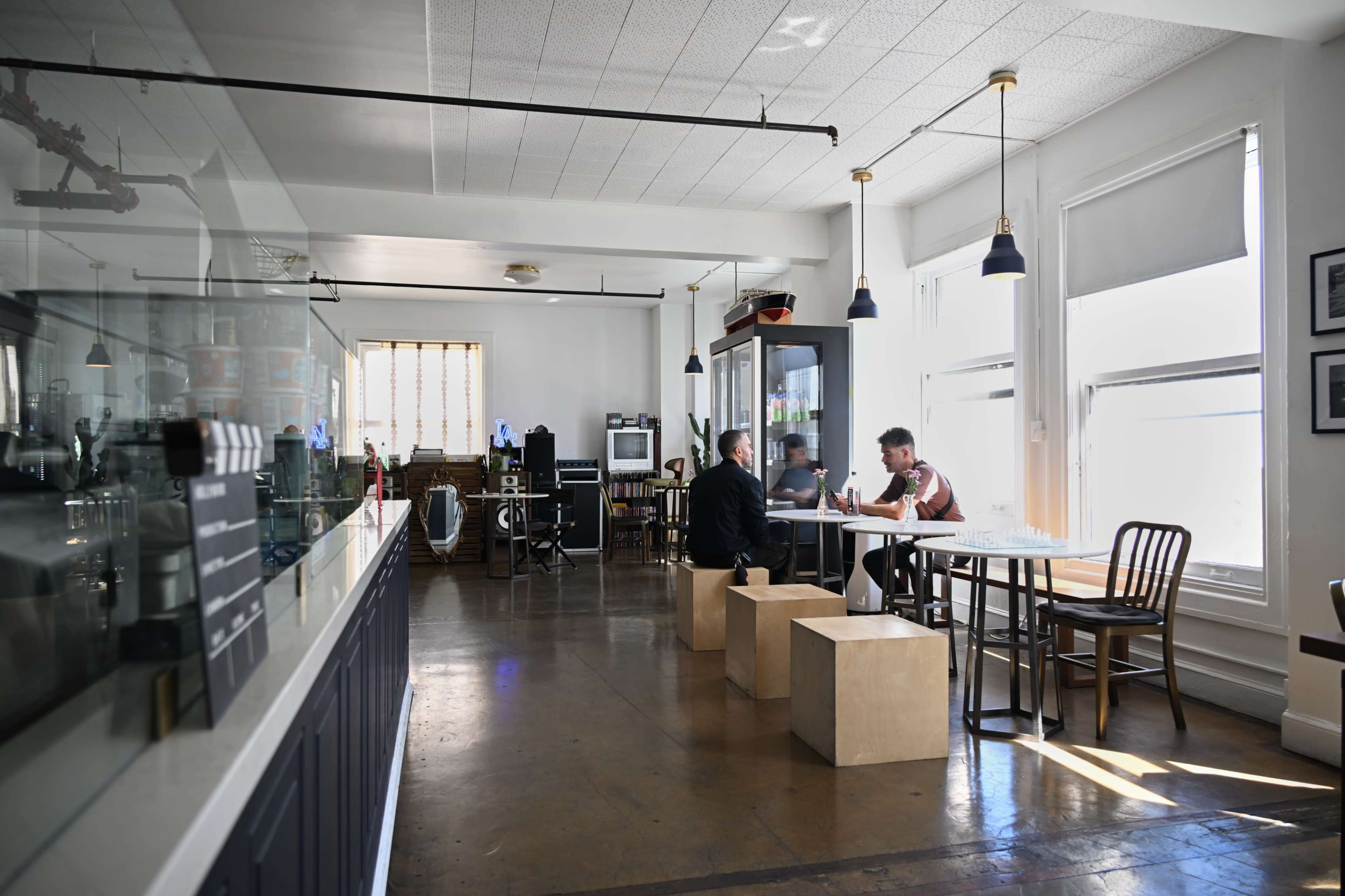 The image shows a modern café interior with a few patrons seated at tables, large windows providing natural light, and minimalist wooden furniture.