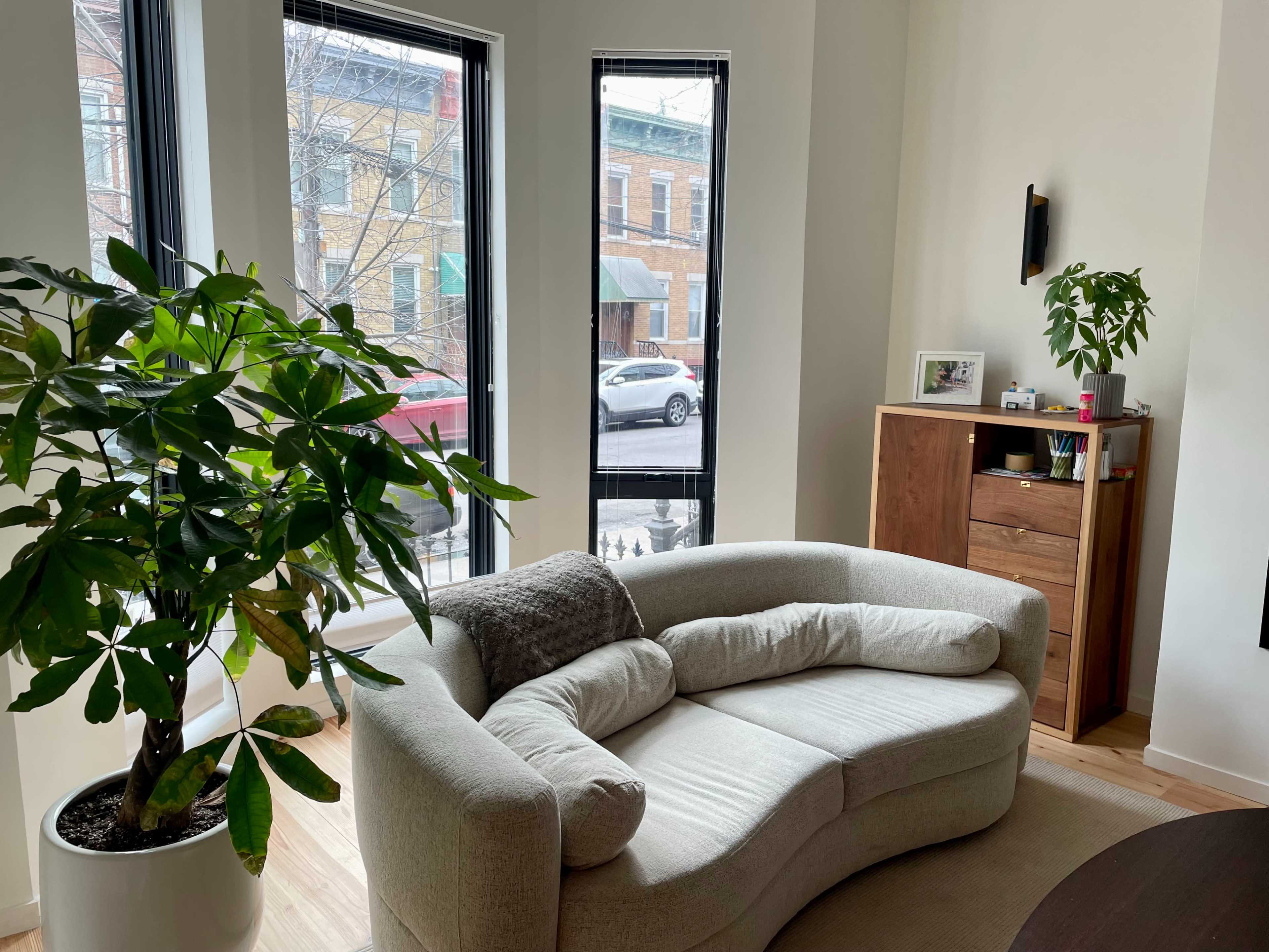 A modern living room features a curved beige couch, a potted plant, and large windows overlooking a street with parked cars.