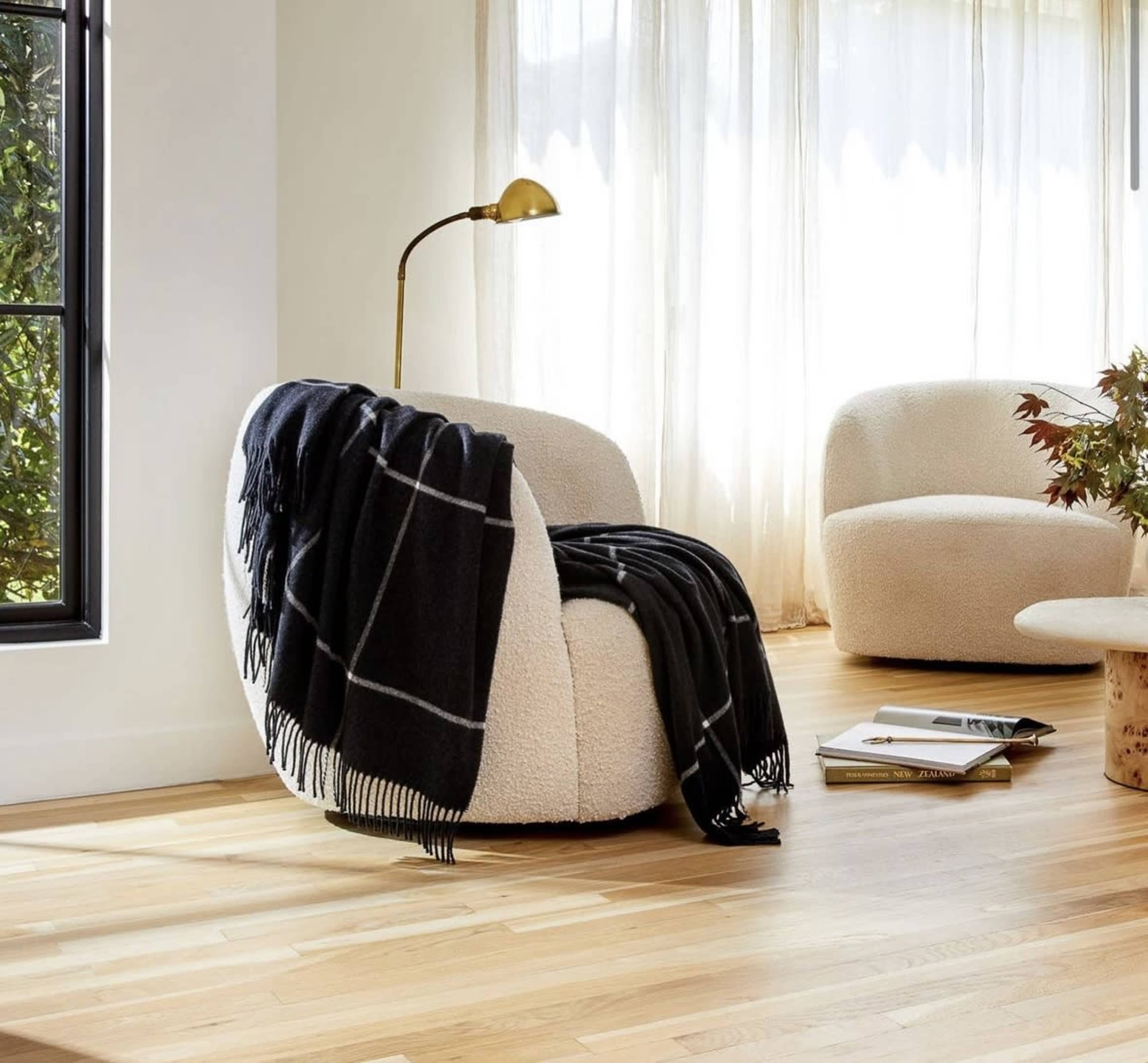 A cozy, rounded armchair draped with a dark blanket sits next to a floor lamp in a sunlit room with large windows and wooden floors.