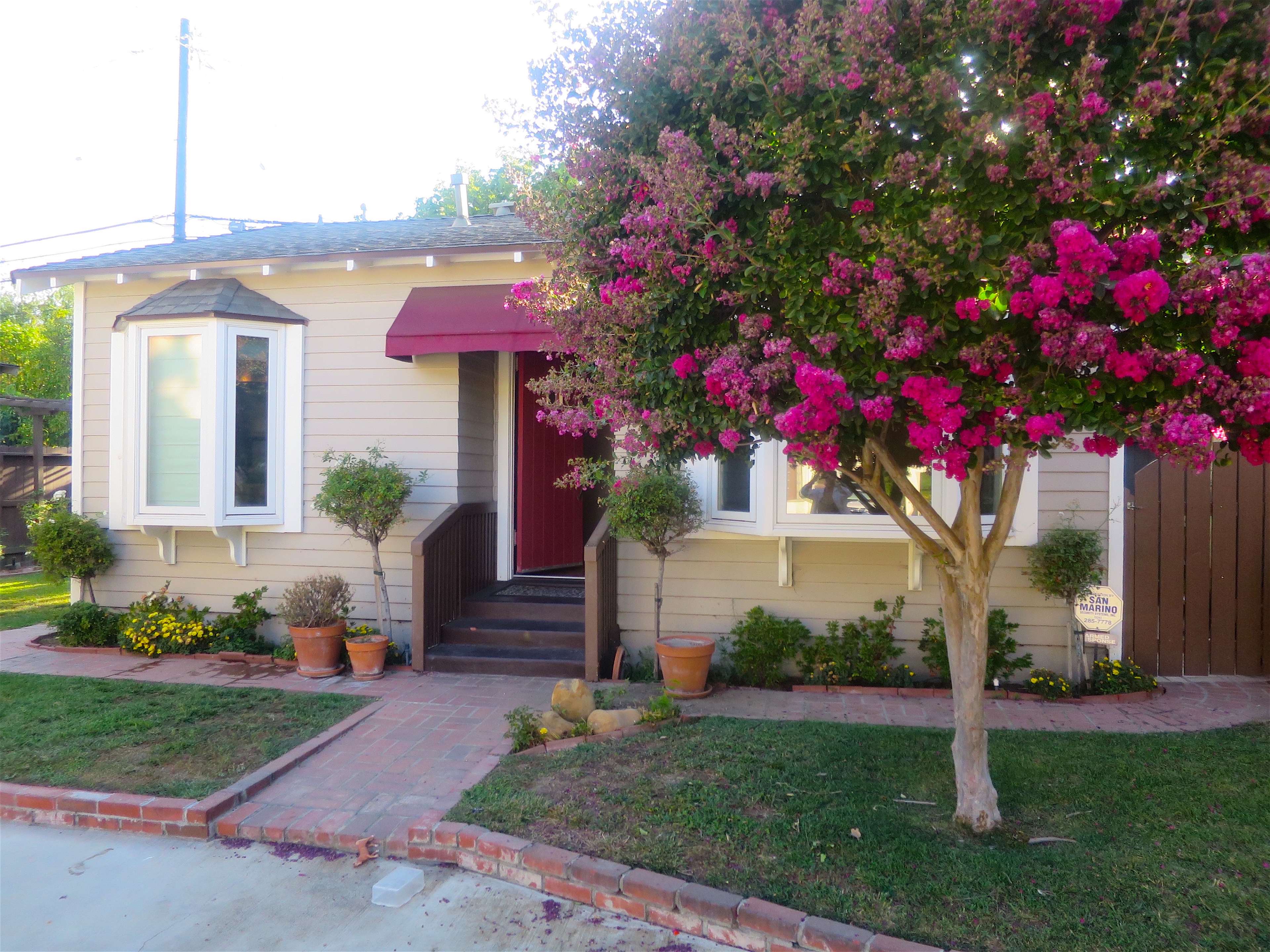 A small house with a red awning is flanked by blooming pink shrubs and potted plants along a brick walkway.