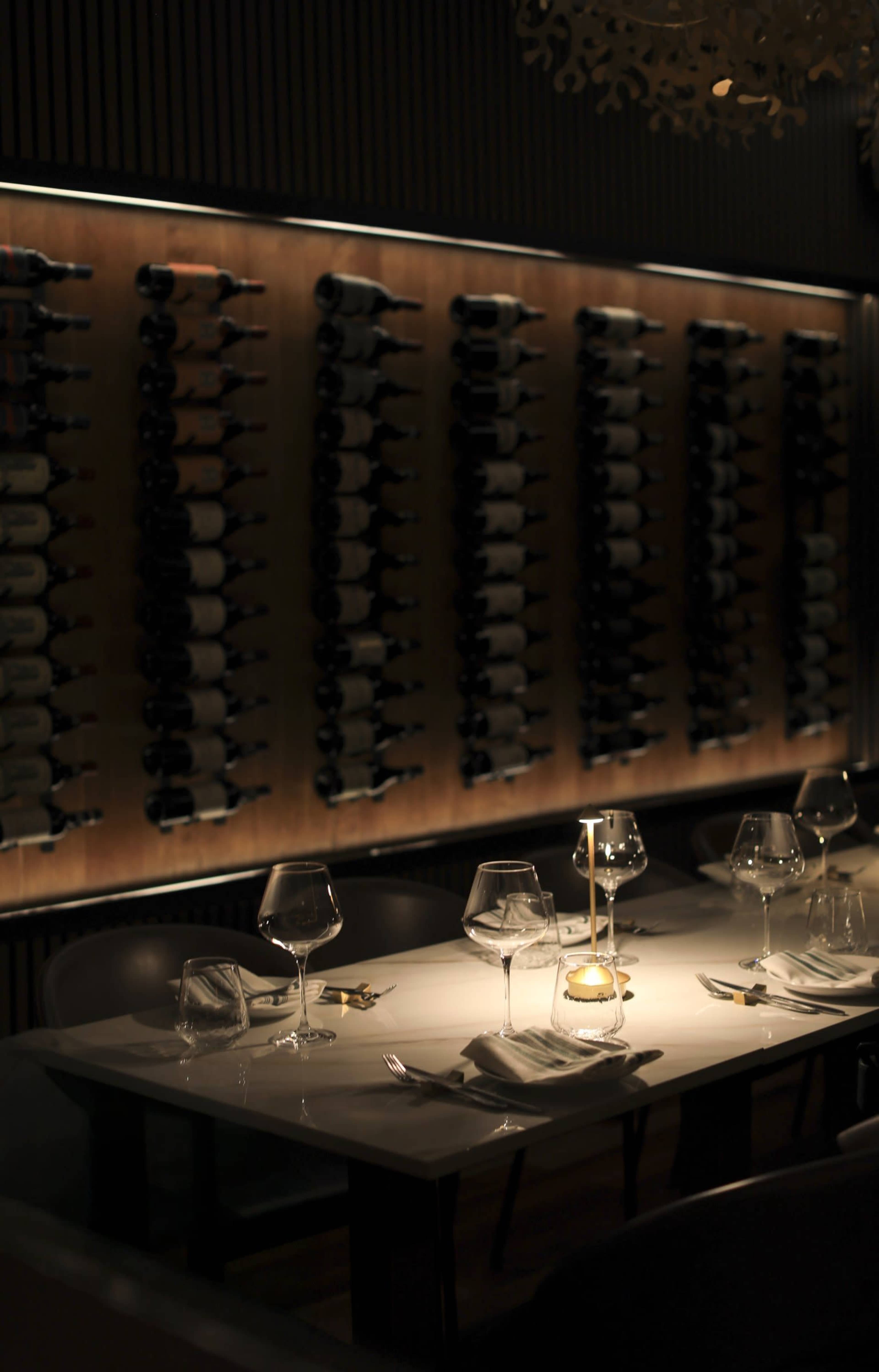 A candlelit dining table is set for several guests, with wine glasses and utensils arranged neatly, against a backdrop of a wall lined with wine bottles.