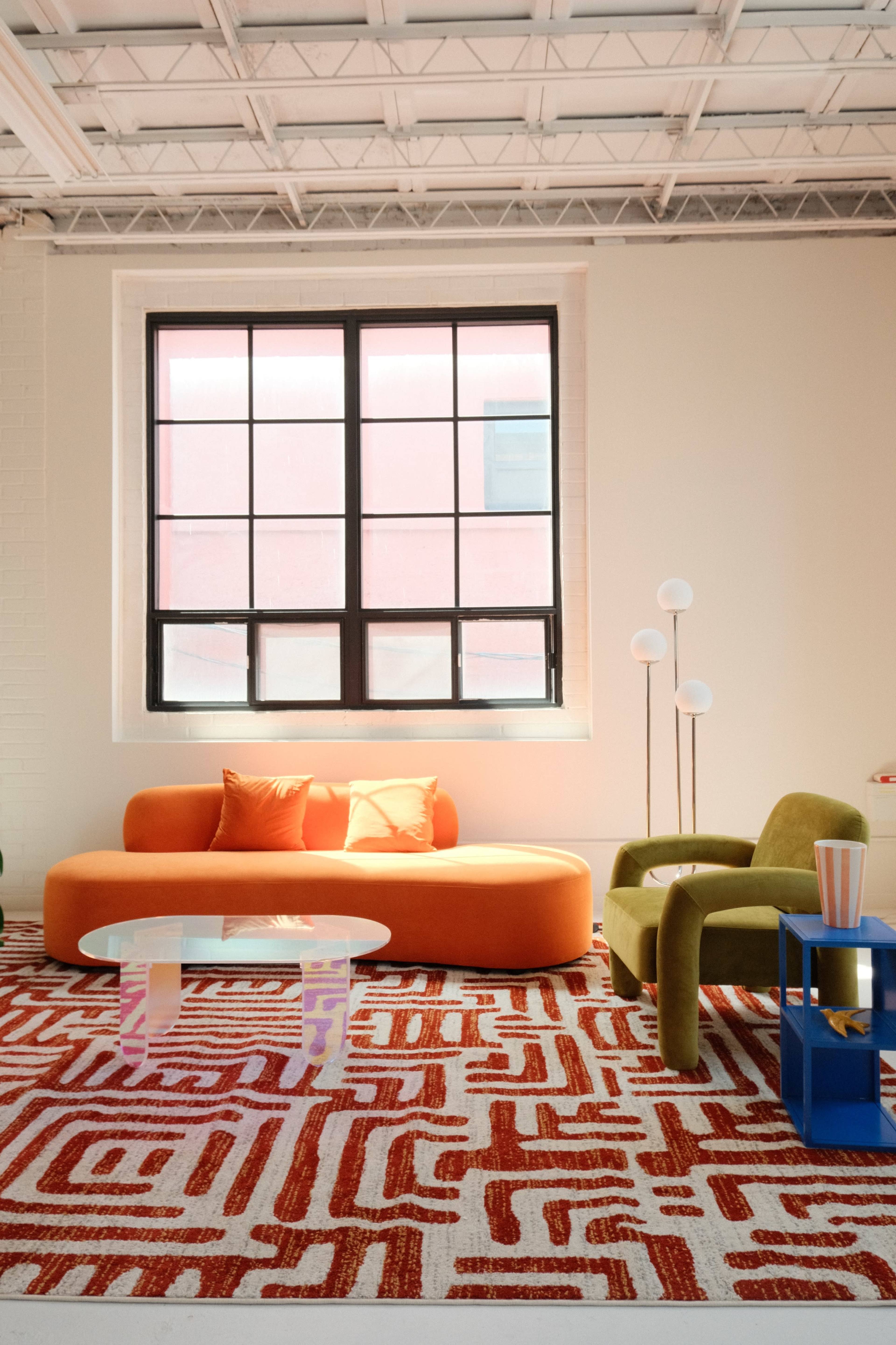A modern living room features an orange curved sofa, a green armchair, a white coffee table, and a patterned red and white rug, with large windows allowing natural light to enter.