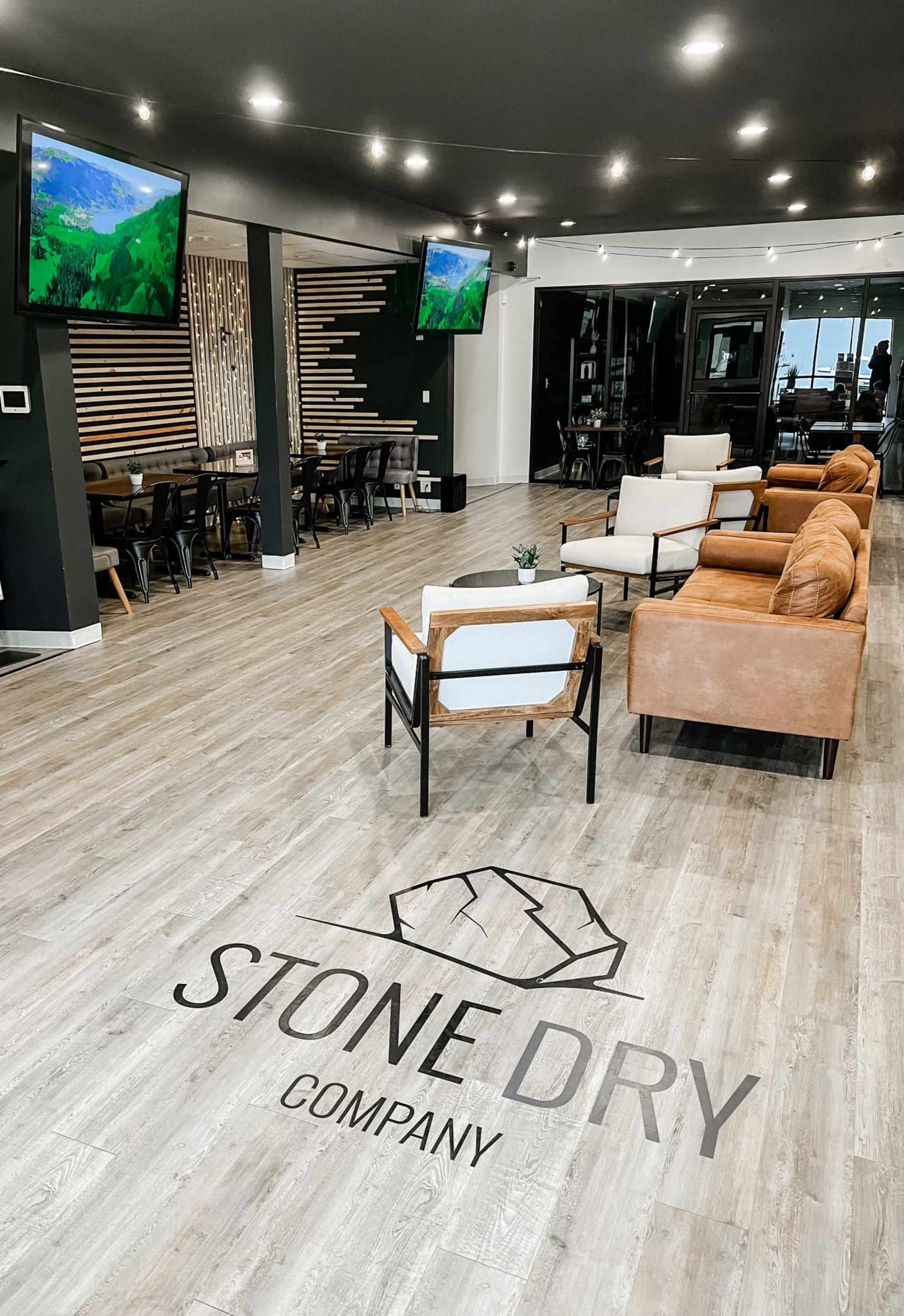 An interior space of a modern office with wooden flooring, lounge seating, and televisions displaying landscapes, featuring a prominent logo that reads "STONE DRY COMPANY" on the floor.