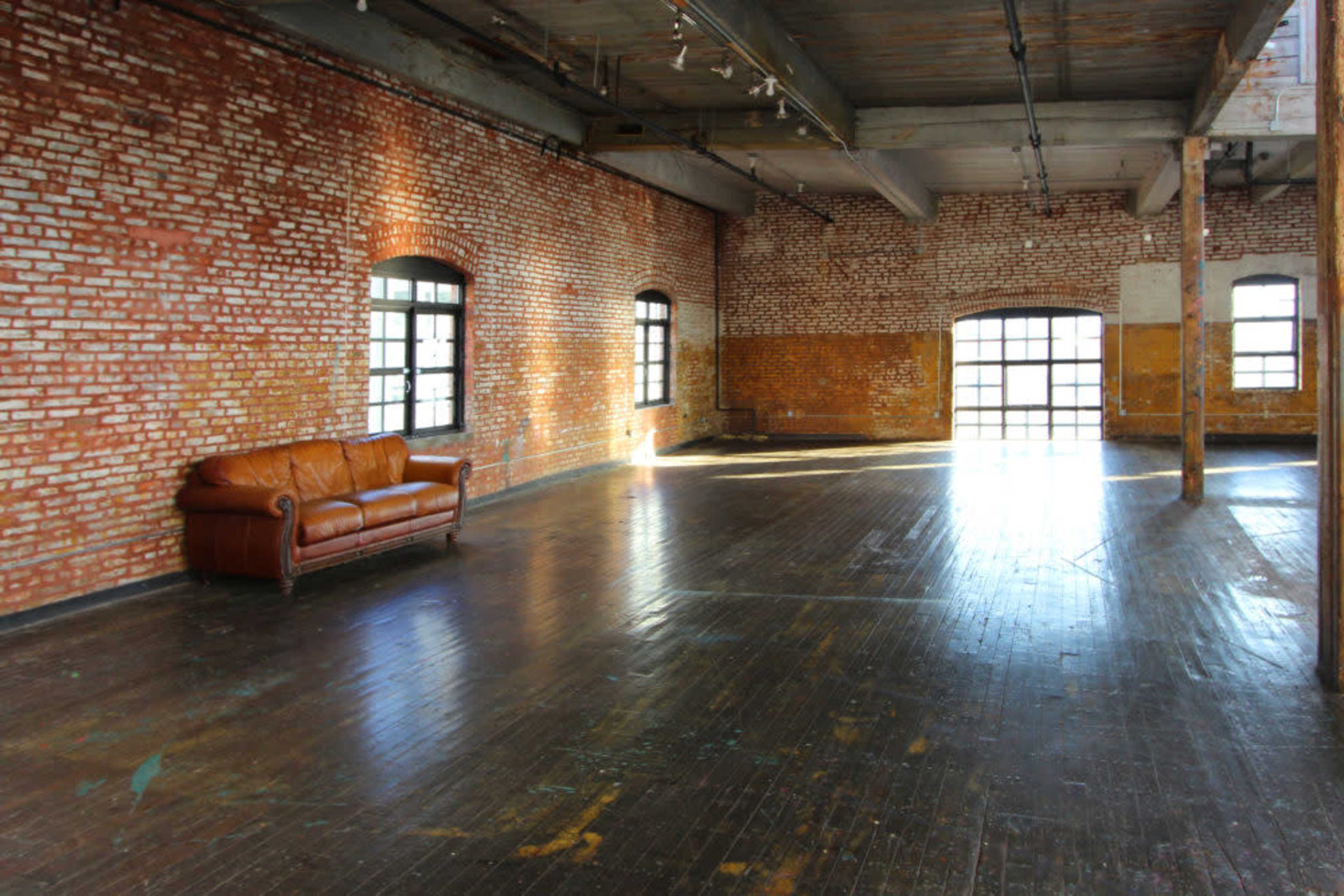 An empty room features exposed brick walls, large windows, and a brown leather couch placed against one side.