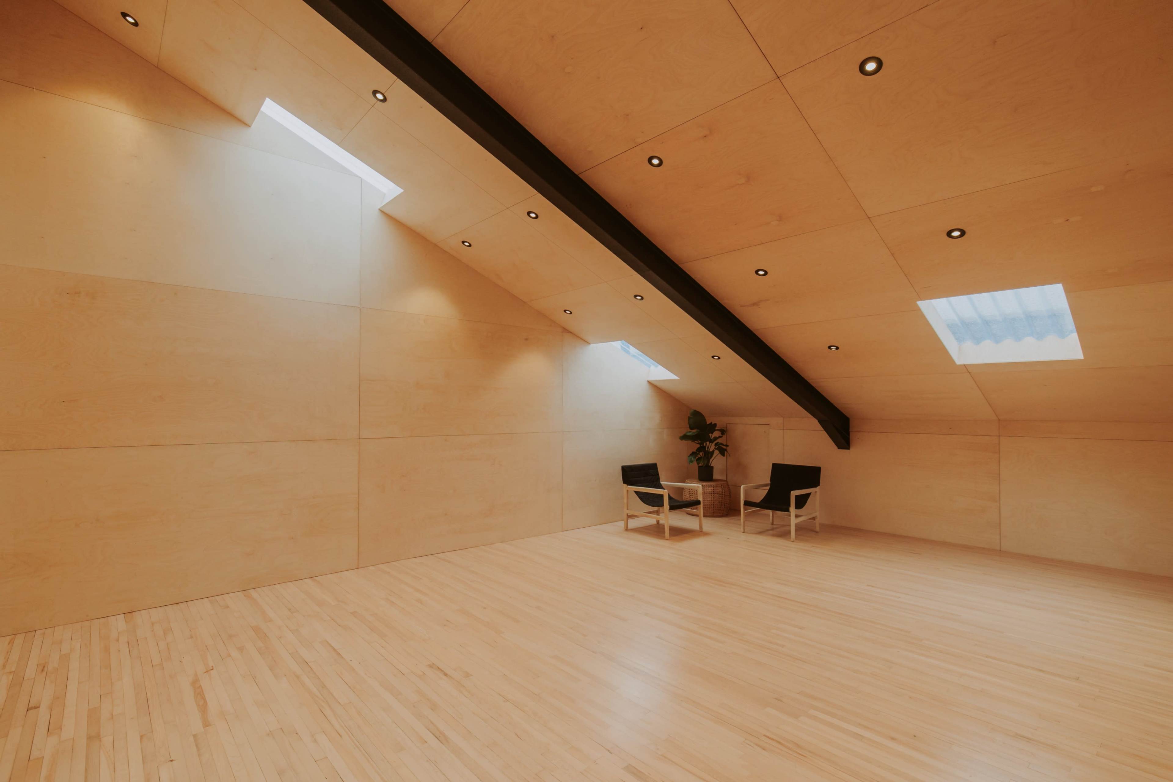 A minimalistic room with wooden walls and flooring, featuring two black chairs and a small table under skylights.