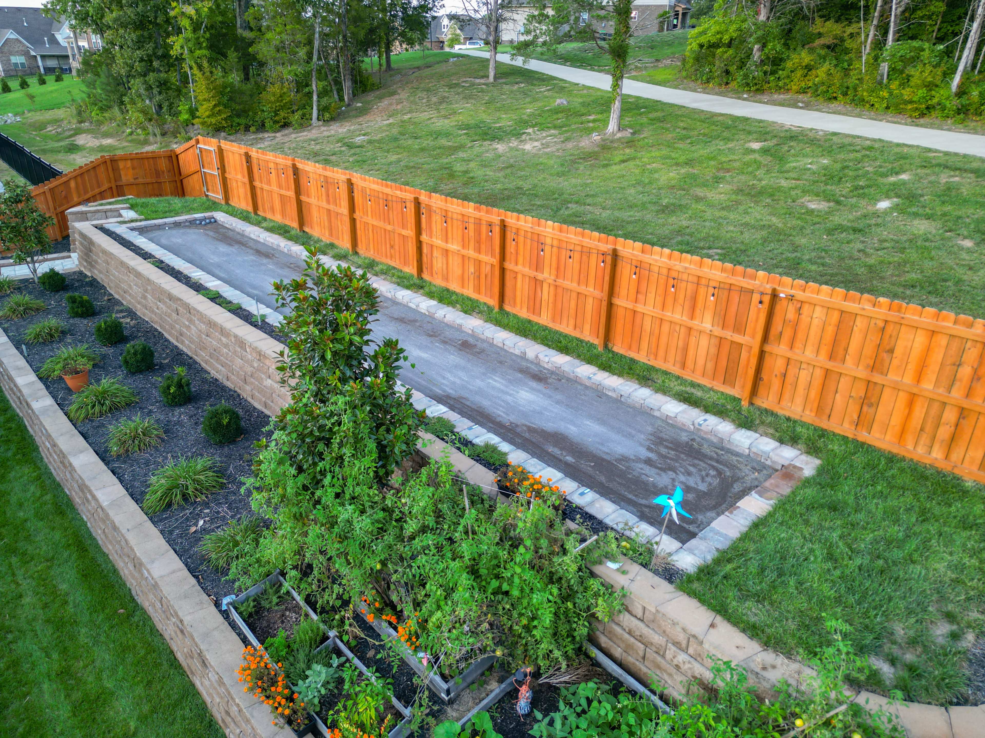 The image shows a fenced backyard featuring a concrete pathway and garden beds with various plants along the edges.