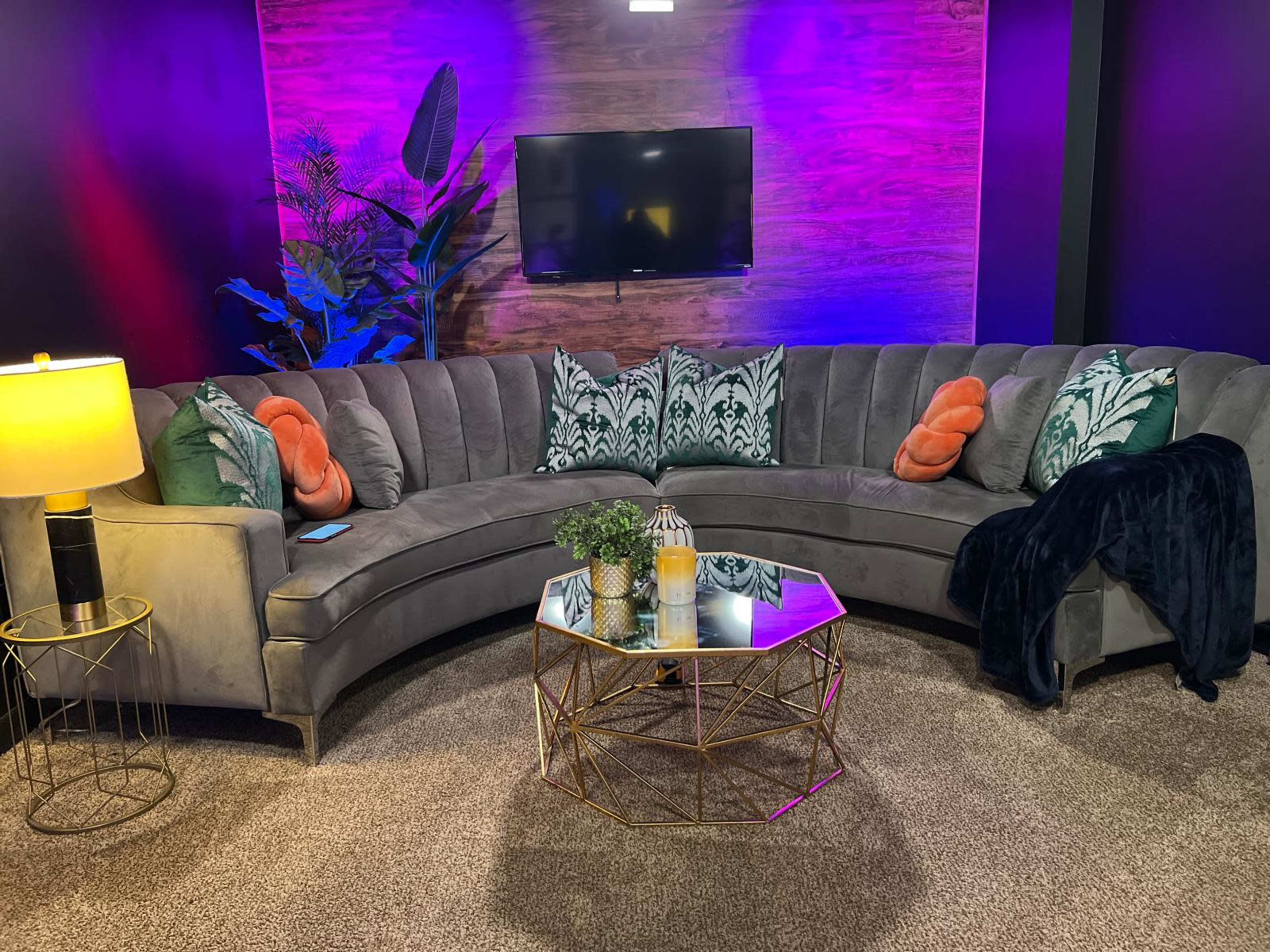 A curved gray sectional sofa with decorative pillows is positioned in front of a wooden accent wall and a television, accompanied by a gold lamp and a geometric coffee table.