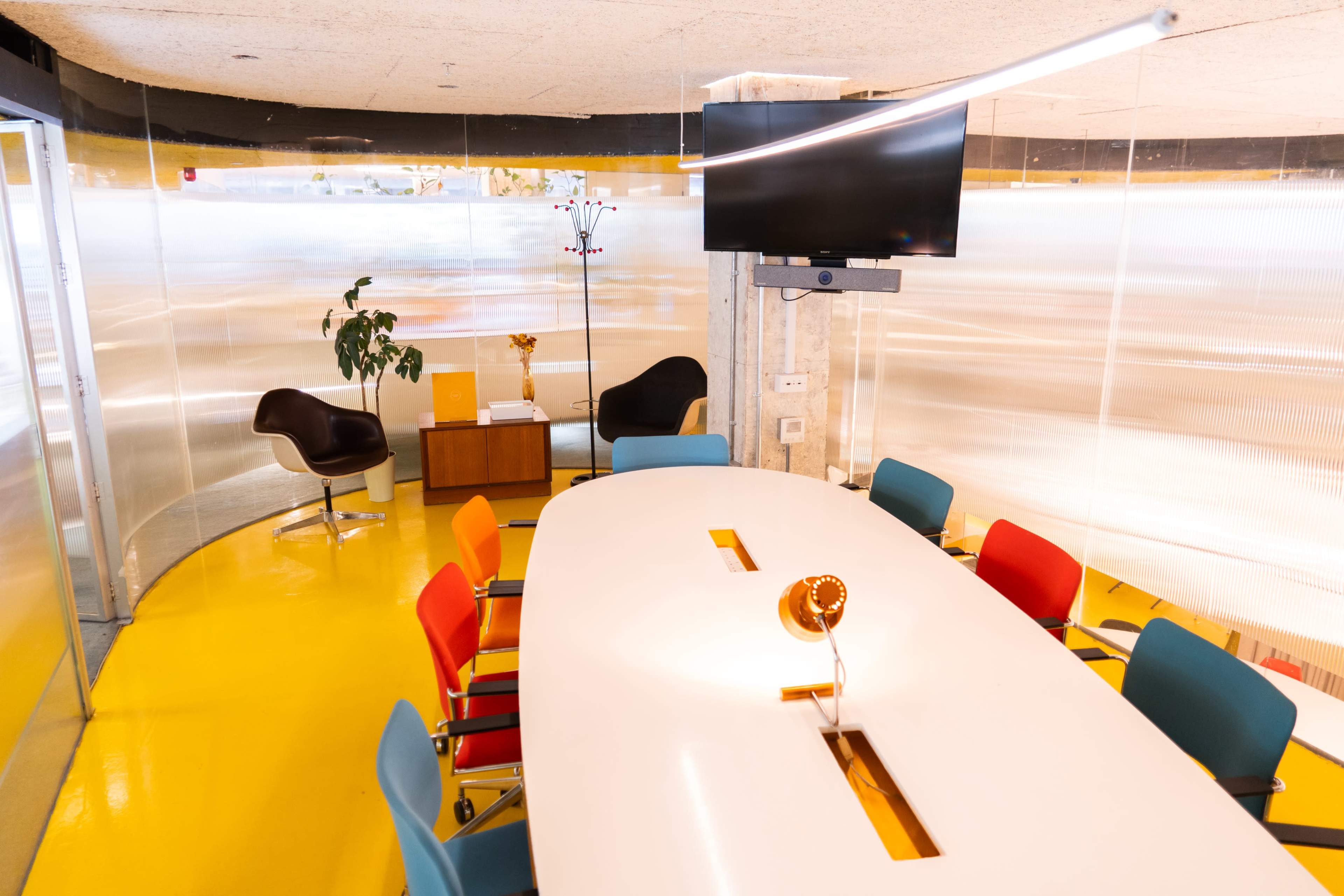 A modern conference room features a large white table surrounded by colorful chairs, with a television mounted on the wall and a plant in the corner.