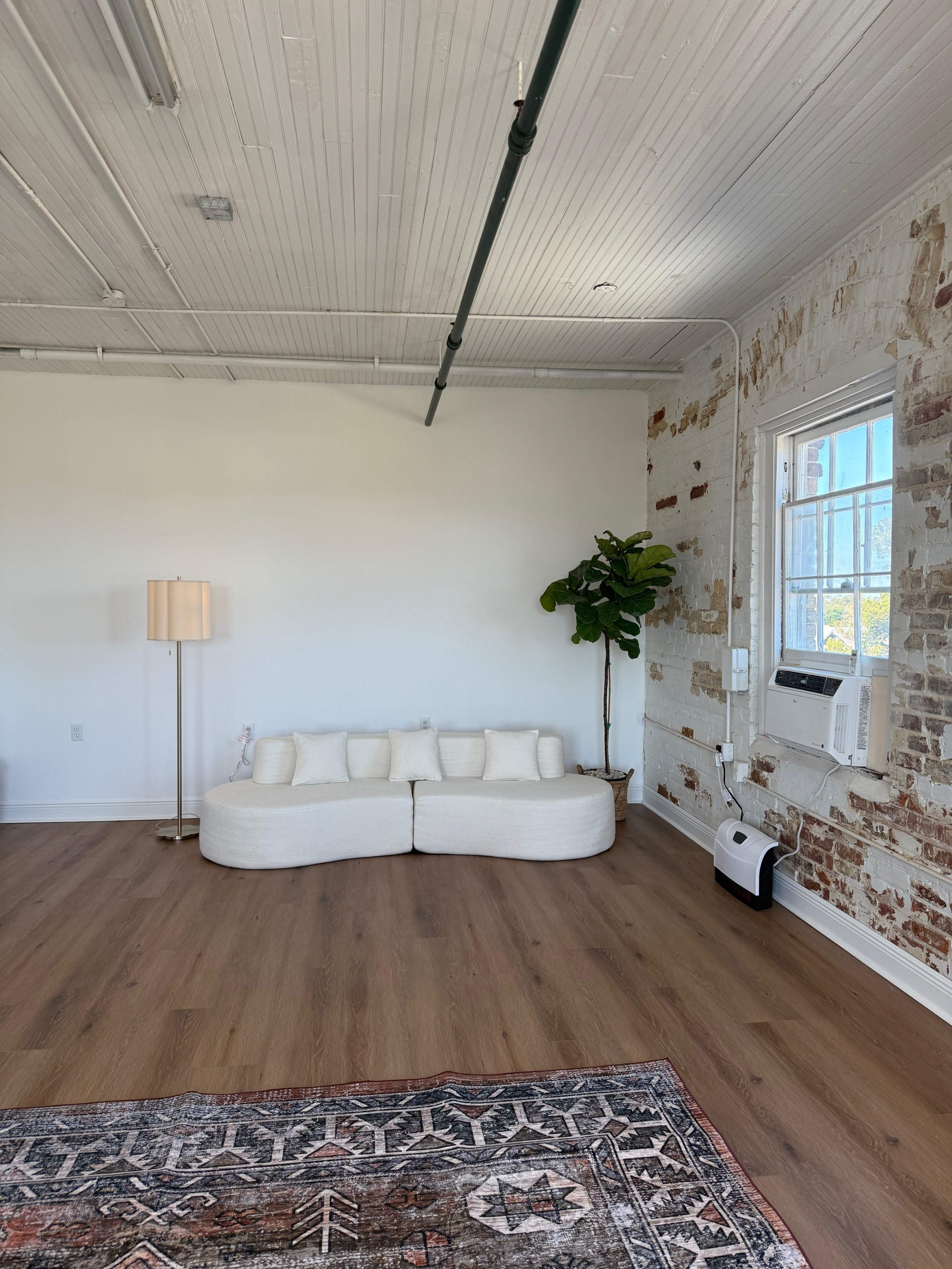 The image shows a minimalist room with a curved white sofa, a floor lamp, a potted plant, and a patterned area rug against a brick wall.