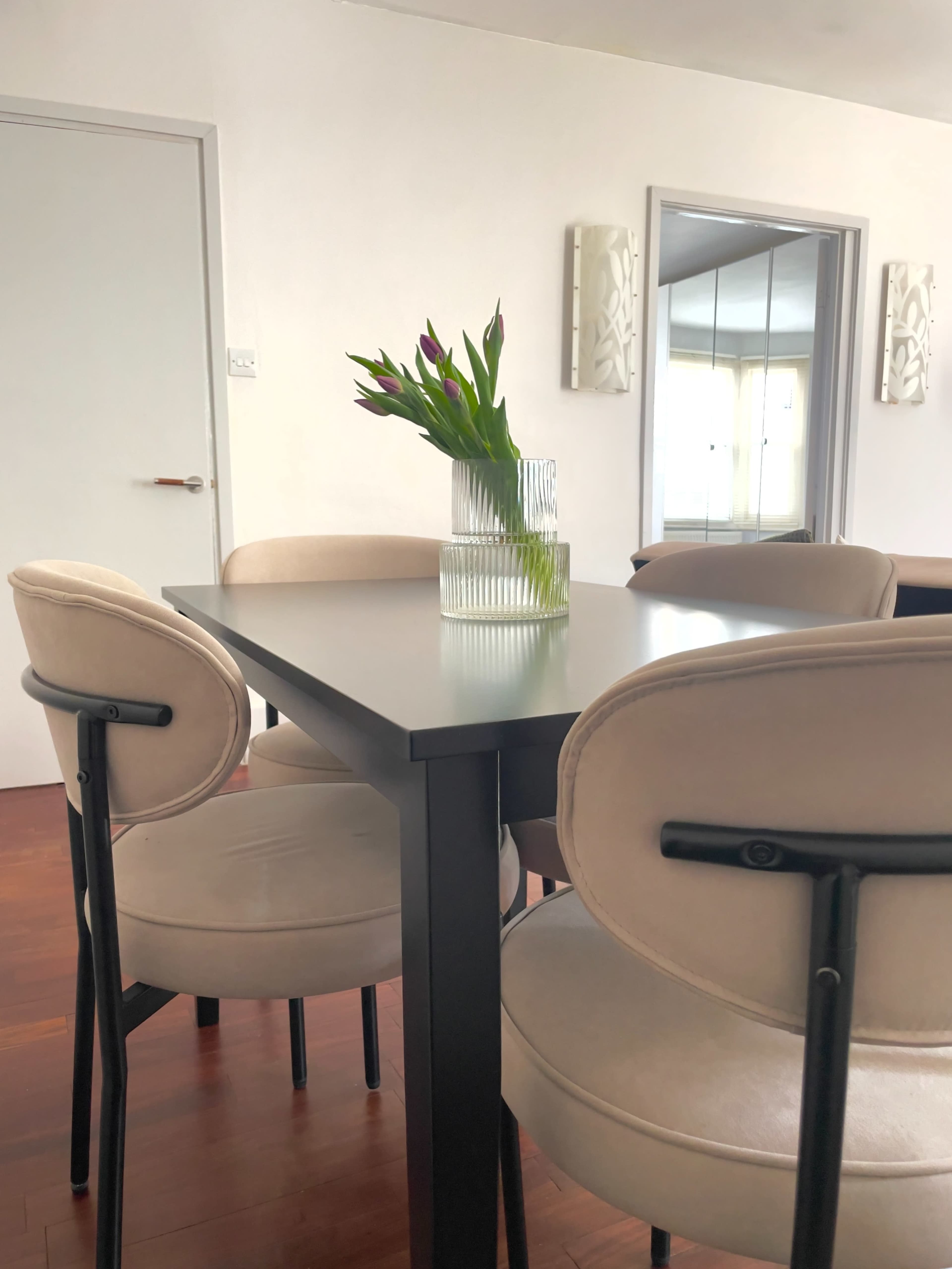 A modern dining table with four upholstered chairs surrounds a vase of tulips in a well-lit room.
