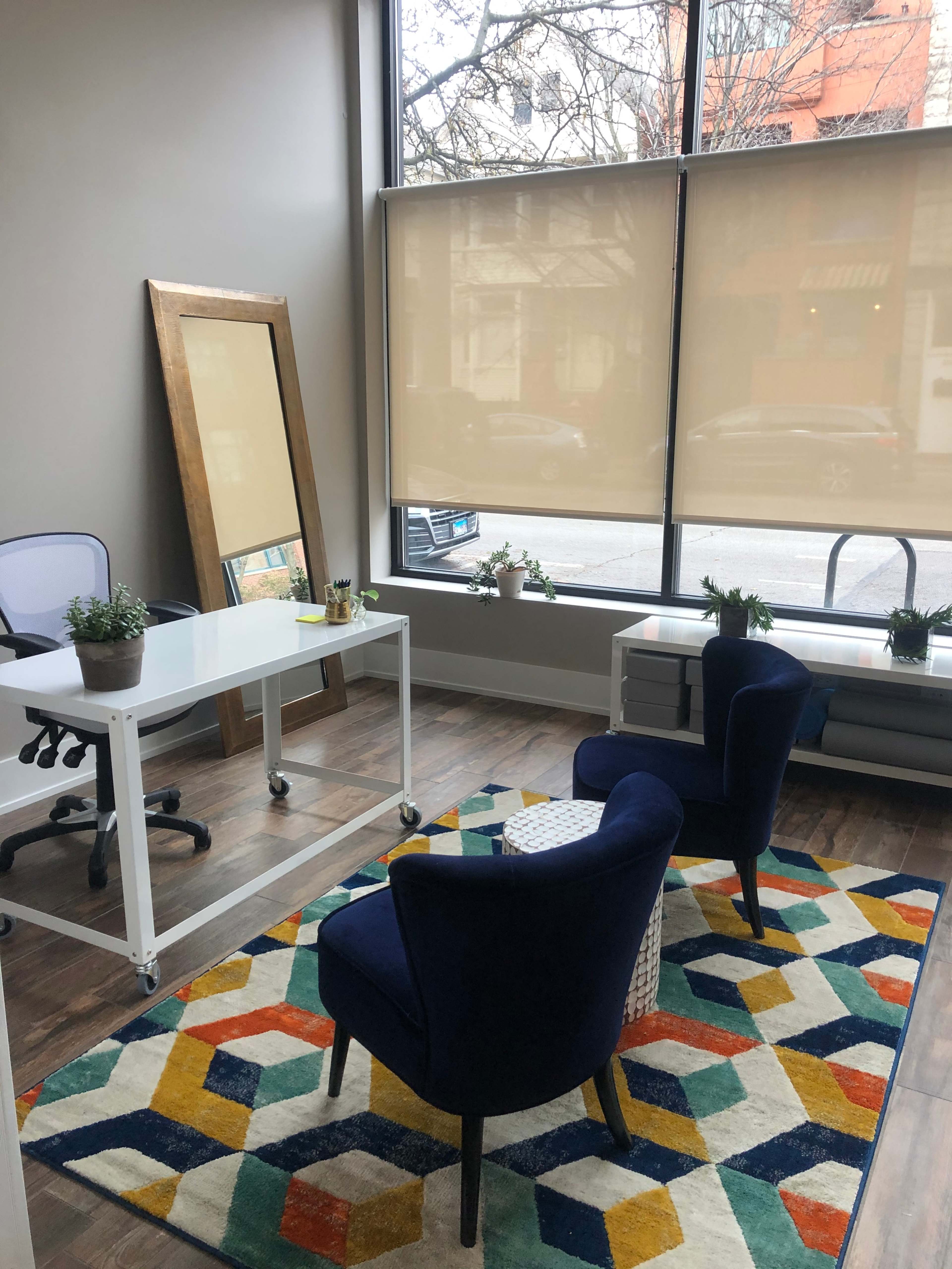 A minimalist office space with a white desk, two blue chairs, and a large mirror, illuminated by natural light from a window with shades.