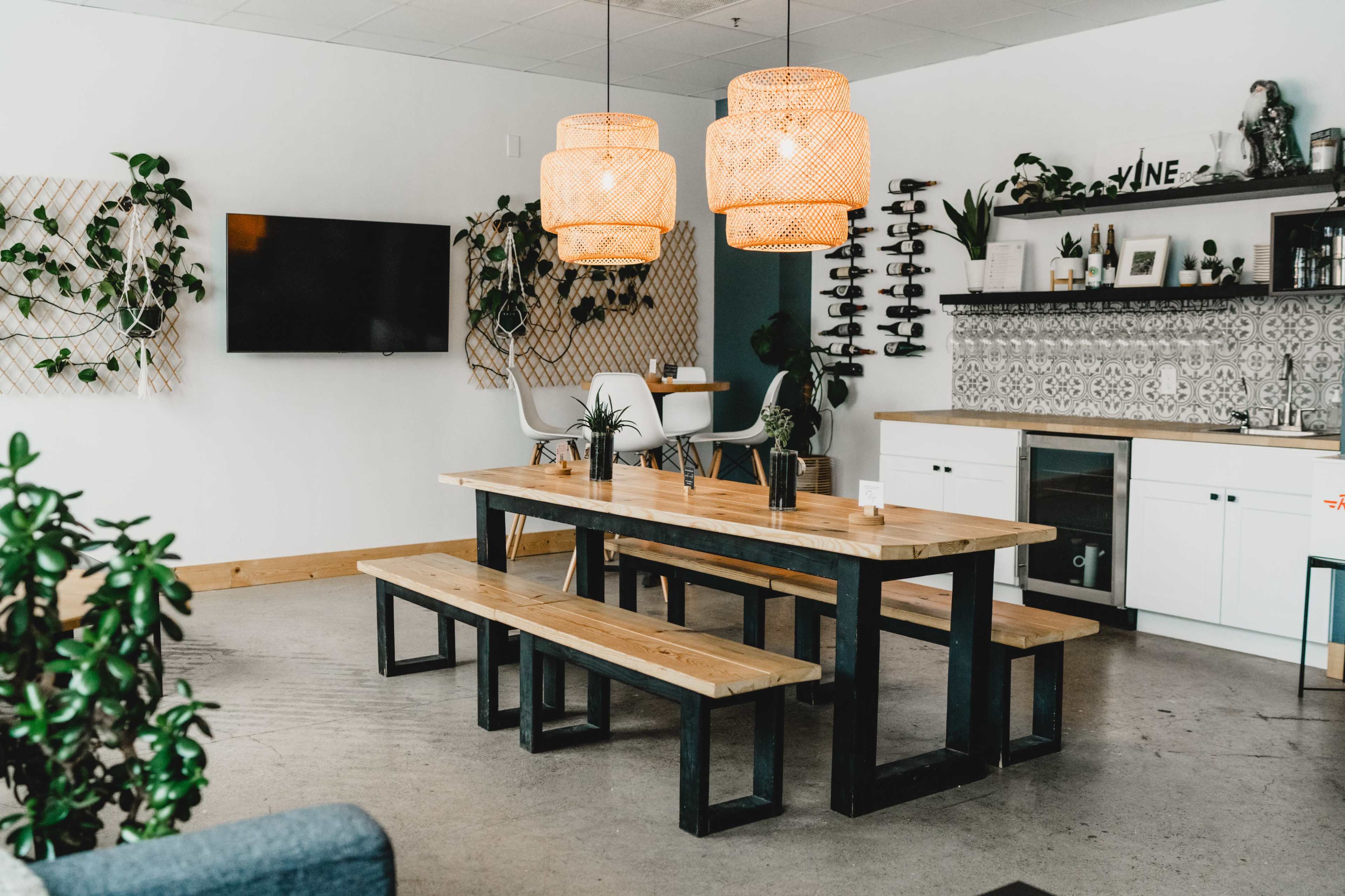 A dining area features a large wooden table with benches, modern light fixtures, and plants, set against a white wall with a television and decorative shelves.