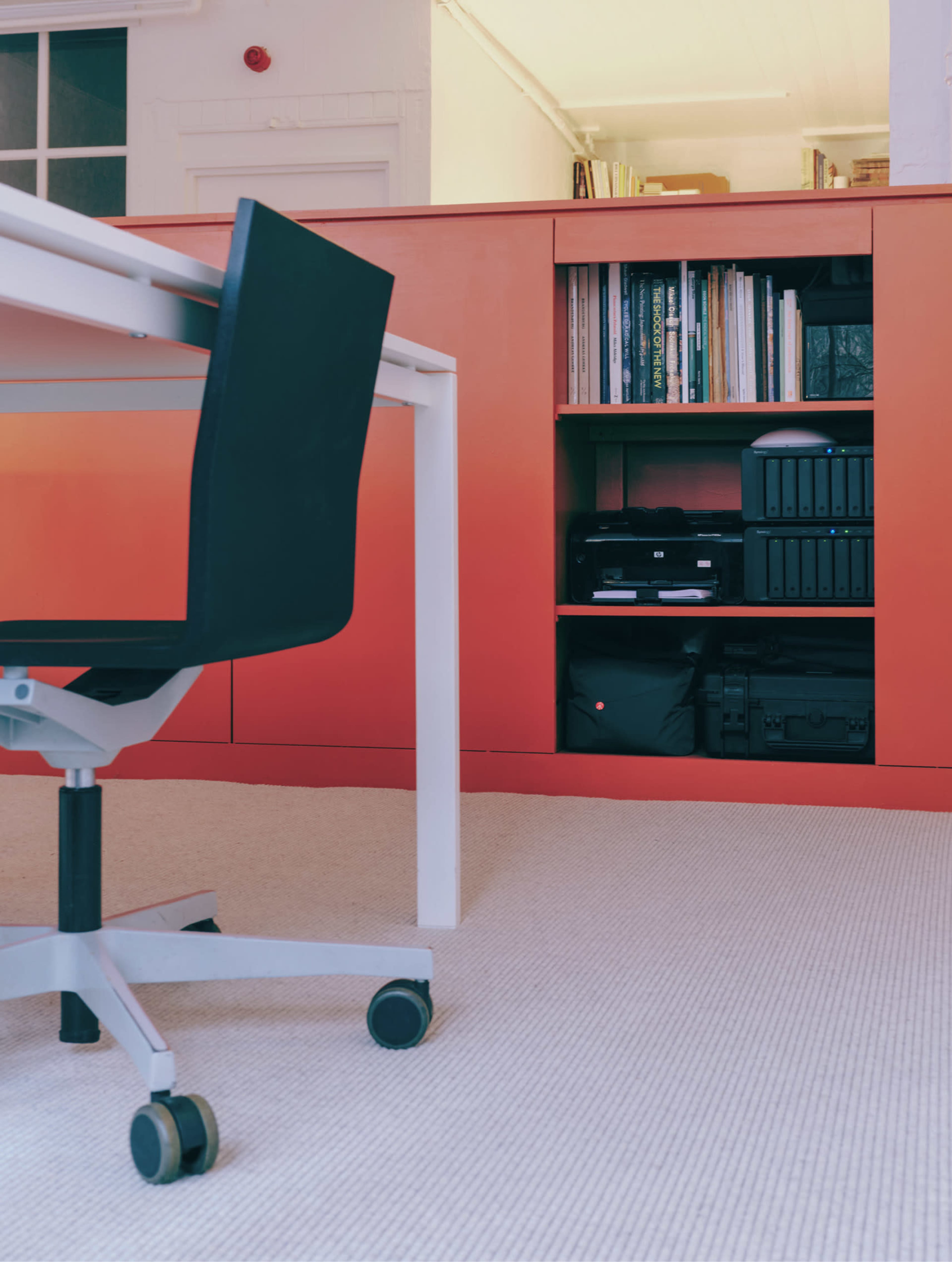 The image shows a modern office space with a white table, a black chair on wheels, and a shelf containing books and equipment against an orange background.