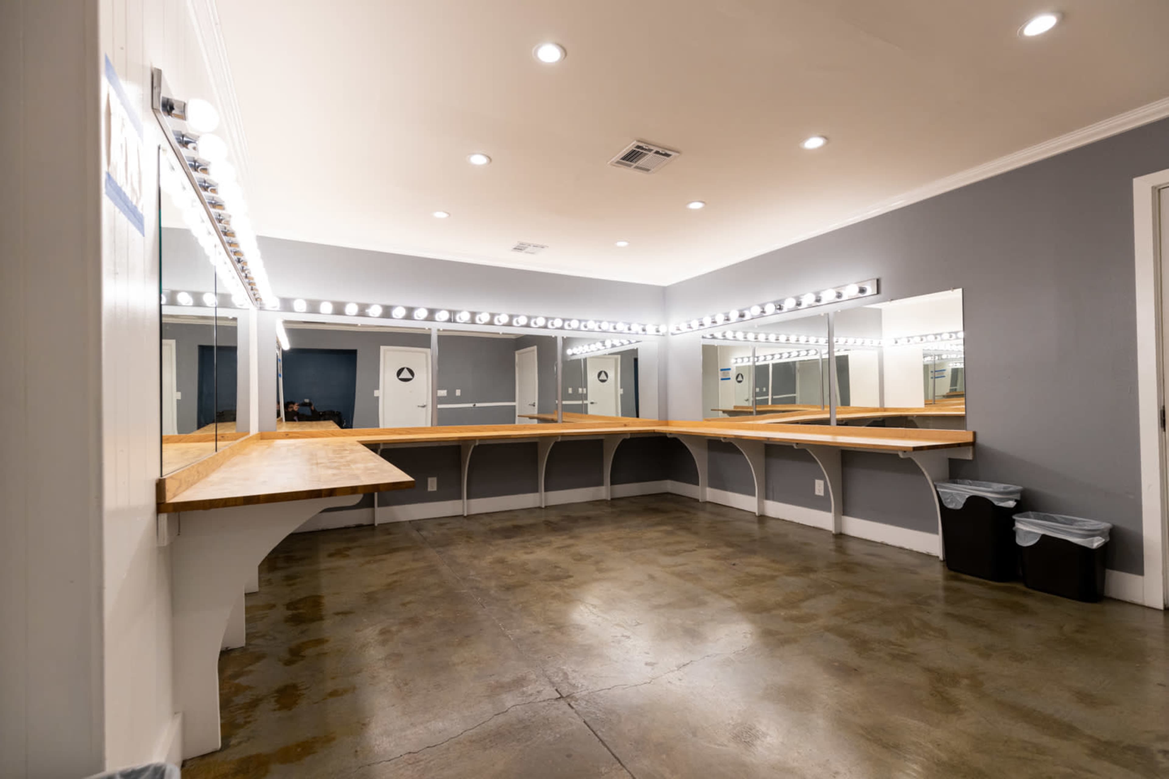 The image shows a spacious dressing room with multiple mirrors and bright lights, featuring a wooden counter and a concrete floor.