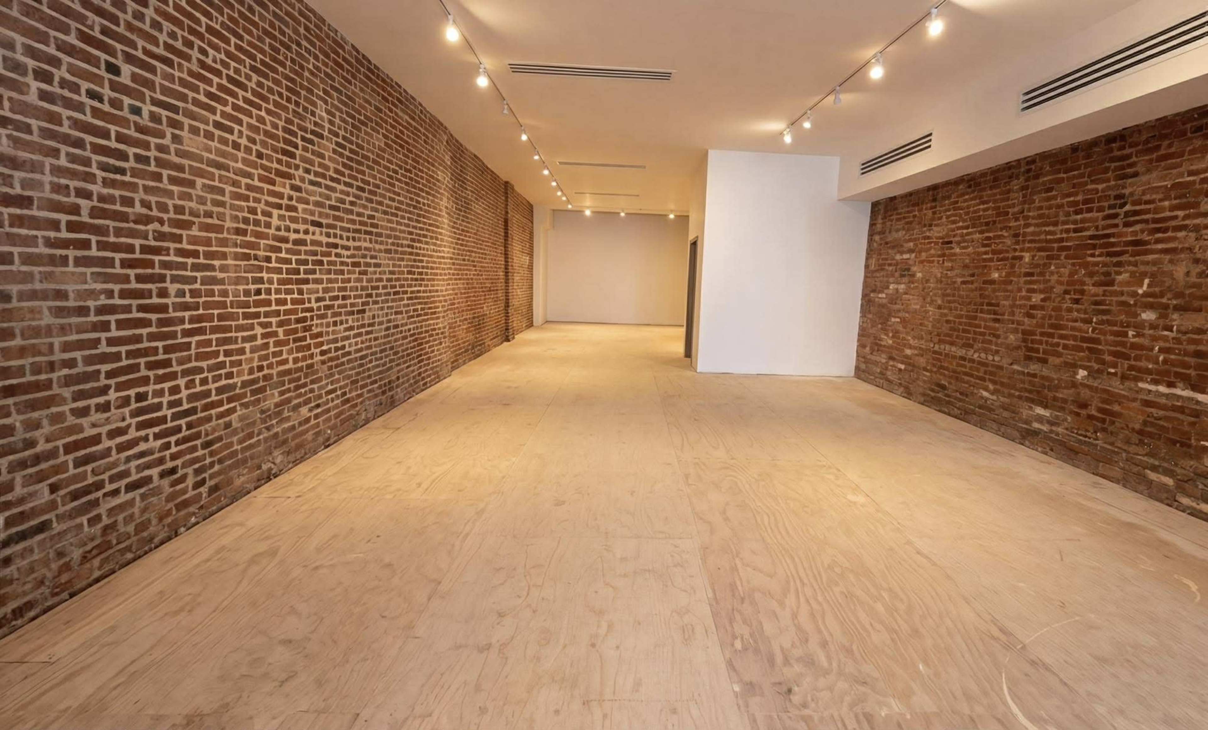 The image shows a spacious interior with exposed brick walls and a wooden floor, illuminated by track lighting along the ceiling.