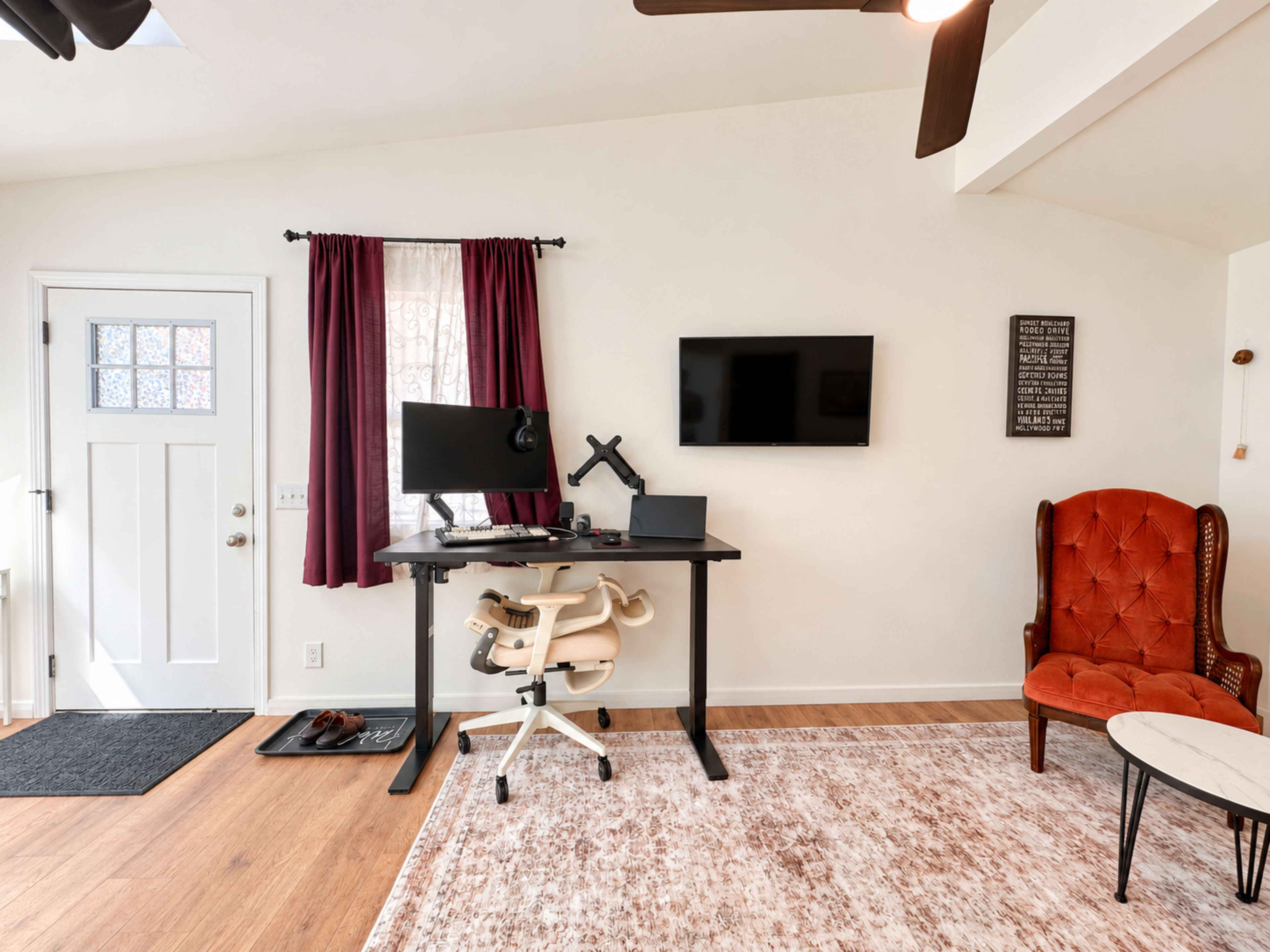 The image shows a minimalistic interior featuring a desk with a computer setup, a television mounted on the wall, a red upholstered chair, a door, and a window with curtains.