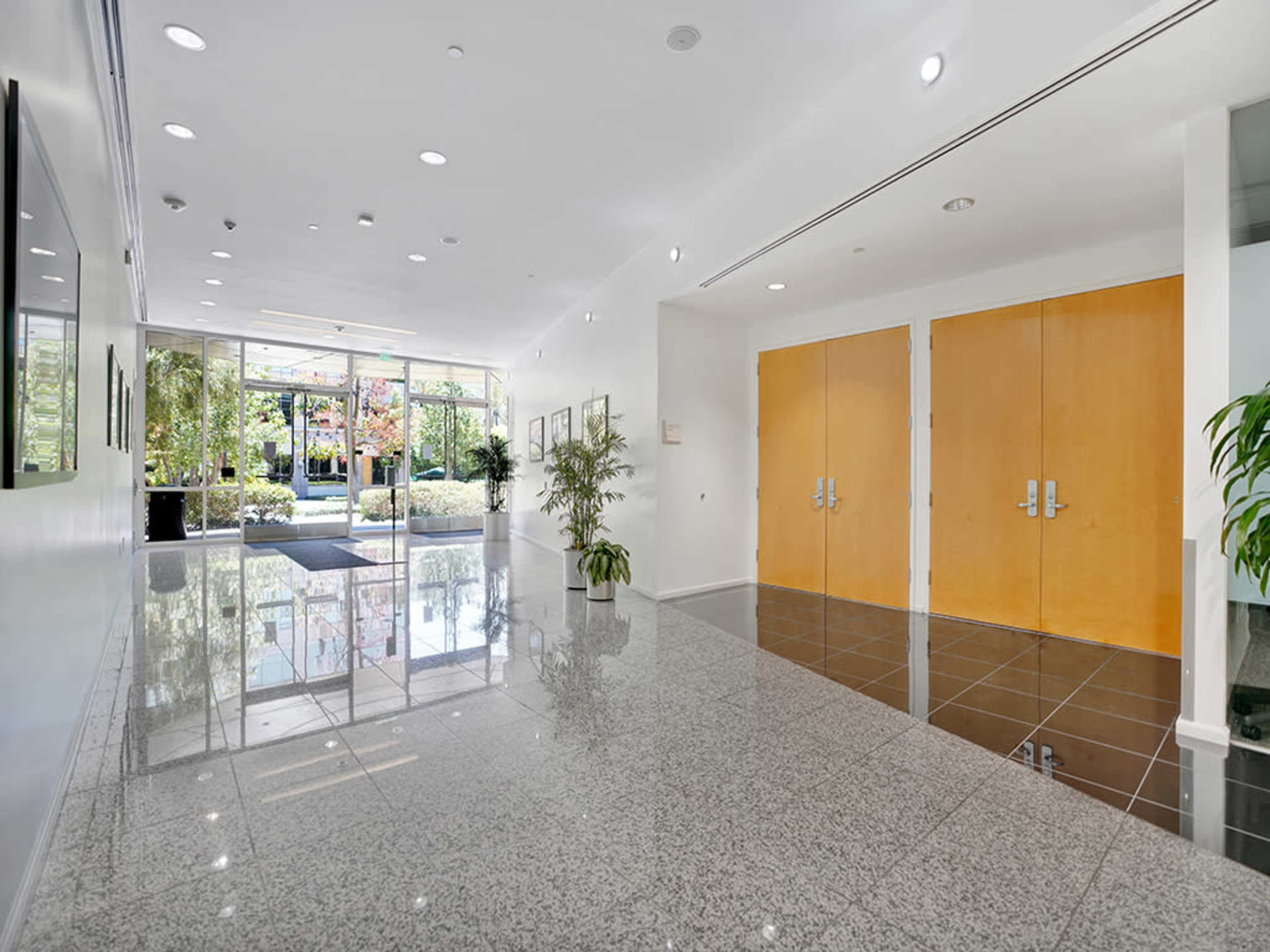 The image shows a spacious, well-lit lobby with glossy floors, large glass doors at the entrance, and greenery on both sides.